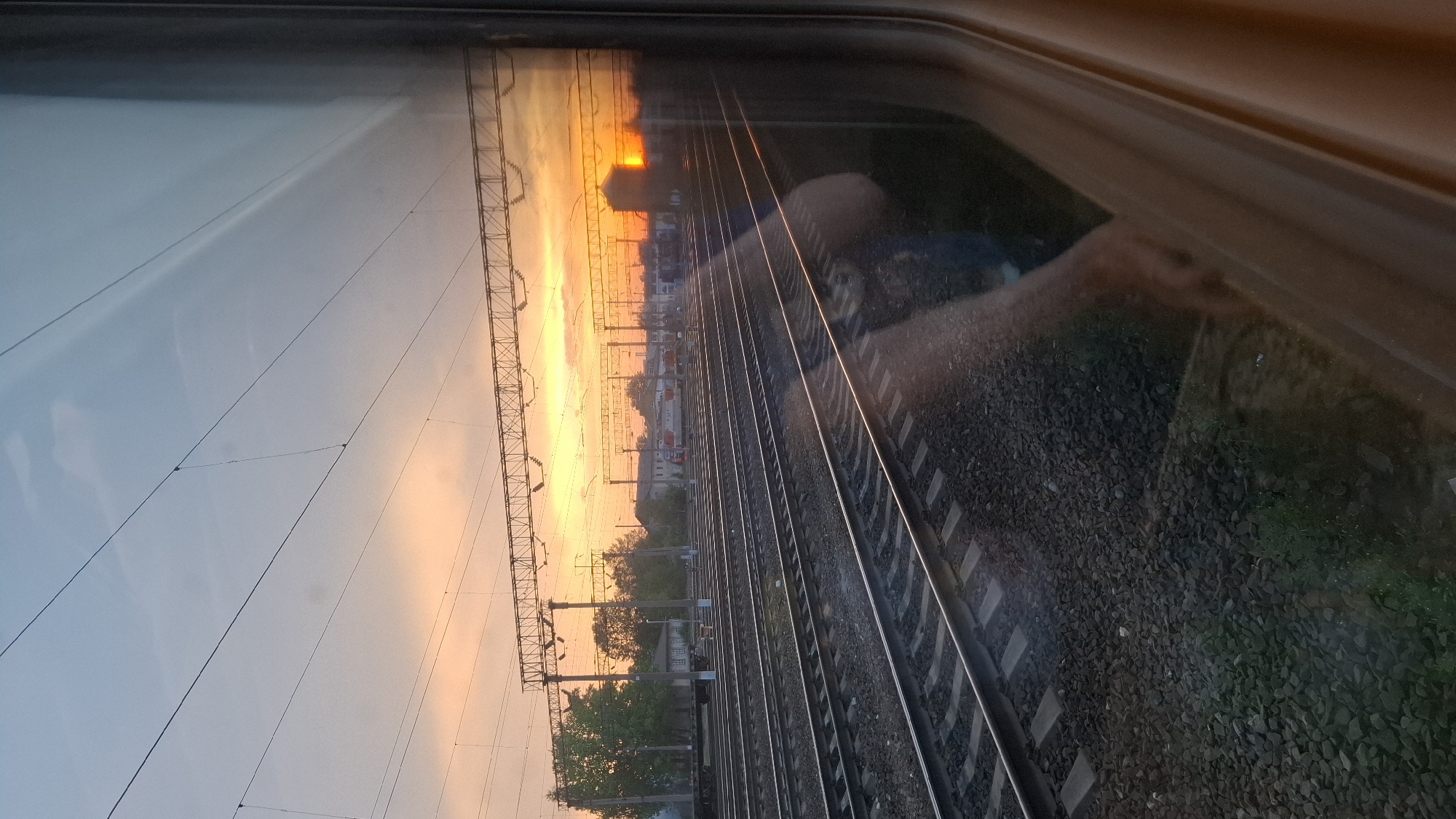 A view from a train window shows multiple parallel railway tracks extending into the distance, reflecting a sunset with hues of orange and yellow sky. The tracks are bordered by ballast. Utility poles and lines run parallel to the tracks. Buildings are visible on the horizon. The reflection of a person is visible in the window. Based on the available context of Buy, Russia, the location is a railway junction in that city.