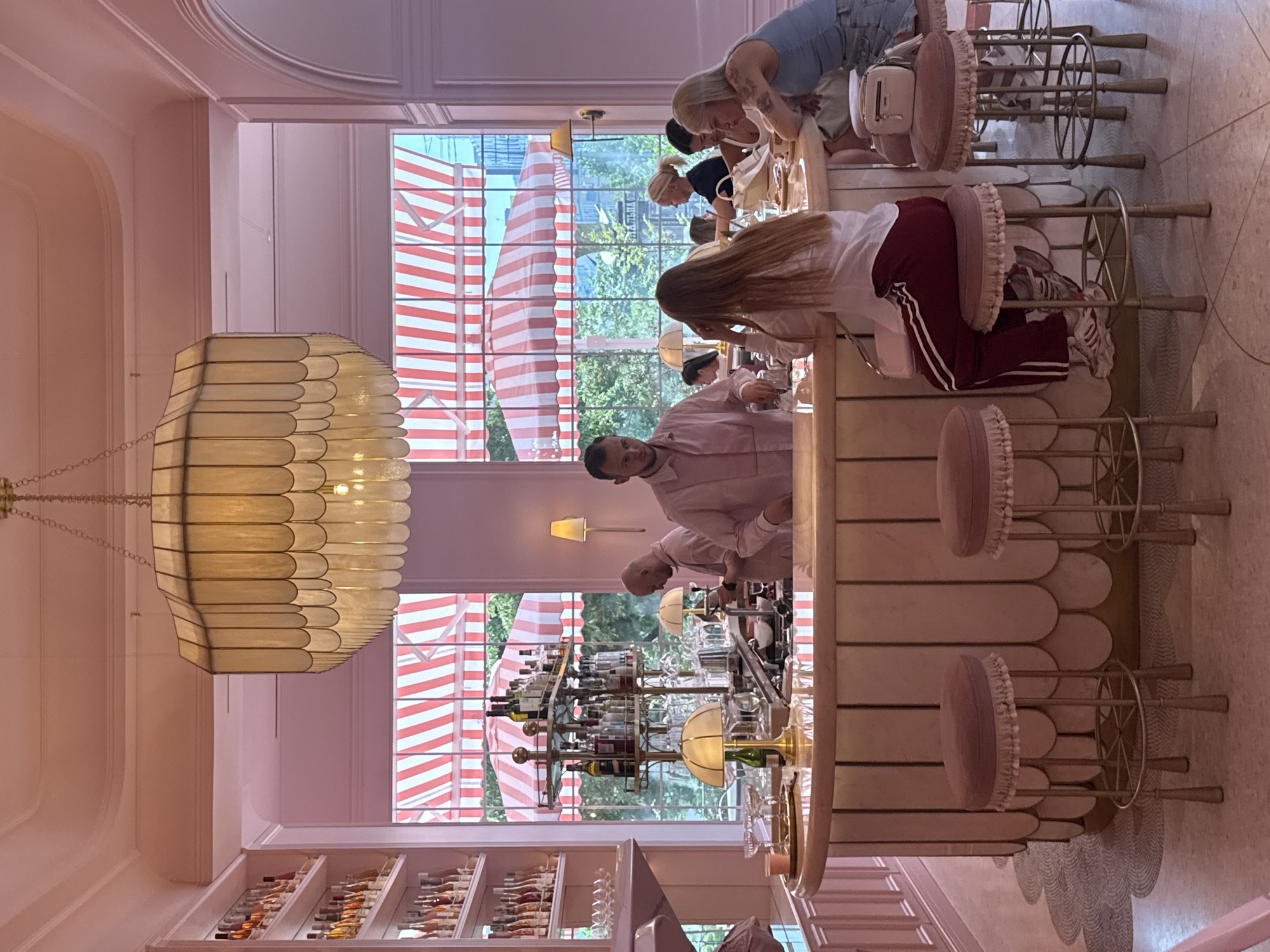 The image depicts the interior of a stylish, pink-themed bar or restaurant. A U-shaped bar with a marble countertop is the central focus, with several patrons seated on plush, pink bar stools. Behind the bar, two bartenders are visible, one in a pink shirt interacting with a customer. A tiered shelf unit behind the bar is stocked with various bottles of alcohol and glassware. Large windows behind the bar reveal an outdoor scene with red and white striped awnings and greenery. The overall aesthetic is modern and chic, with elegant lighting fixtures and decorative shelving. The location is identified as Kyiv, Ukraine.