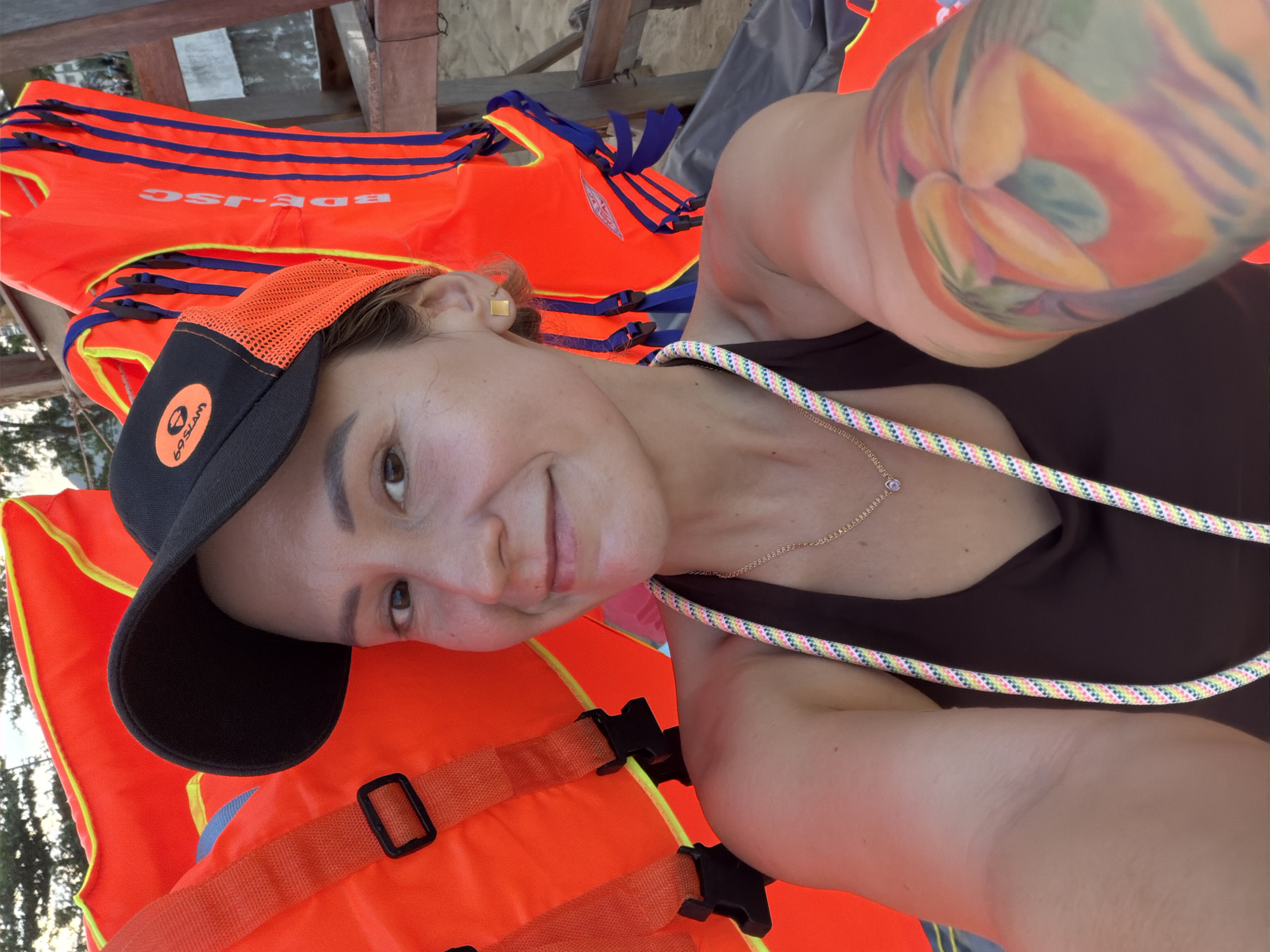 A woman is taking a selfie, lying down on several bright orange life vests. She is wearing a black baseball cap with an orange mesh brim and a circular logo on the front. She is also wearing a dark top, and a delicate gold chain with a small pendant is visible around her neck. A colorful rope is draped across her chest and shoulder. Her left arm, which is visible in the frame, has a large, colorful tattoo of what appears to be fruit or flowers.
The life vests are stacked and folded, with some showing blue trim and yellow accents. Text is visible on one of the life vests, appearing to read "BDE-JSC". The background suggests an outdoor setting, possibly near water, with hints of trees and wooden structures visible behind the life vests. The lighting suggests it is daytime. The woman's expression is relaxed and pleasant. The overall impression is a casual, perhaps preparatory moment before or after an activity involving the life vests, likely near a body of water. The location context is South Quy Nhon Ward, Vietnam.