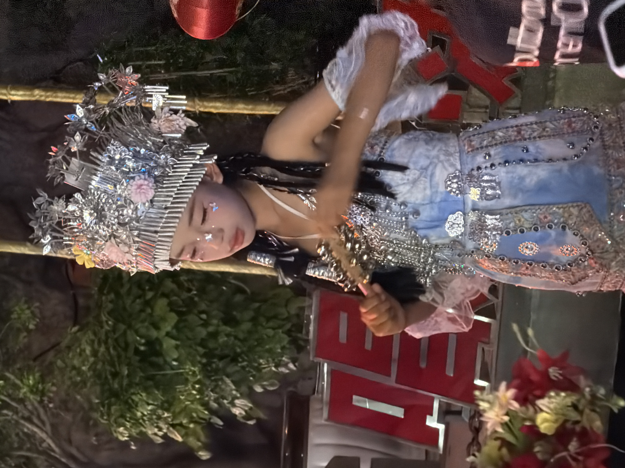 A young East Asian girl, appearing to be of Chinese descent, is depicted in the Yuzhong District of China, likely at an outdoor event or performance. She is adorned in an elaborate, traditional costume, featuring a highly decorative headdress with intricate silver metallic elements, small pink and yellow flowers, and dangling silver chains. Her face is painted with subtle glitter and small butterfly decals on her cheeks and forehead. She is wearing a blue and white garment that appears to be a dress or costume, embellished with shimmering beads, sequins, and floral patterns. A delicate, sheer white fabric is attached to her sleeves. In her right hand, she holds a small, ornate object, possibly a prop or part of her costume. Behind her, red signage with white Chinese characters is visible. The background consists of lush green foliage and a dark, indeterminate setting, suggesting an evening or dimly lit outdoor environment. To the right, a portion of a red lantern is visible. The overall impression is that of a cultural performance or celebration.