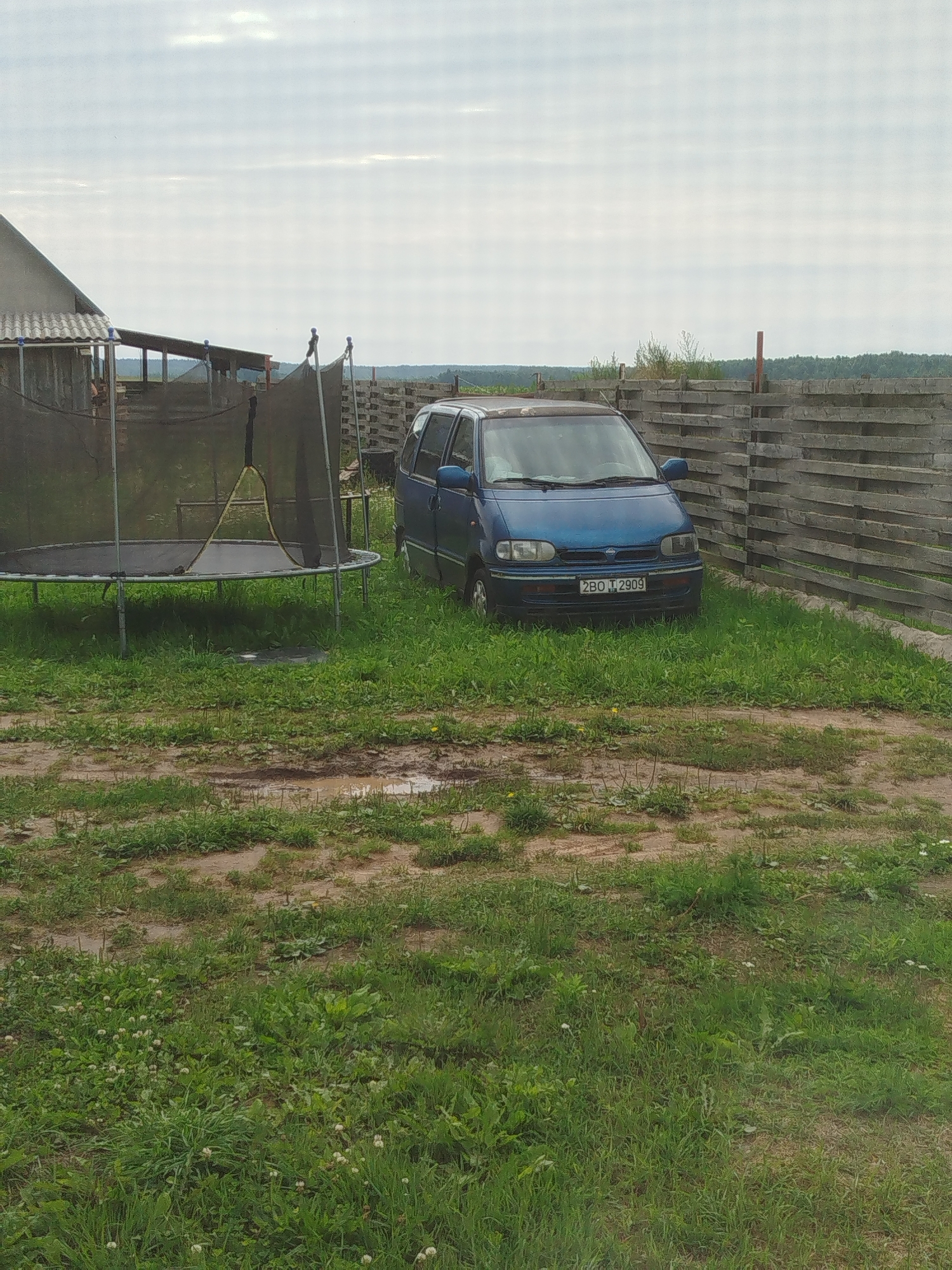 A blue minivan is parked on a gravel and grass area next to a wooden fence. To the left of the minivan is a large trampoline with a black safety net. A building with a sloped roof is visible behind the trampoline. The sky is overcast. The terrain appears uneven with patches of mud and scattered vegetation.