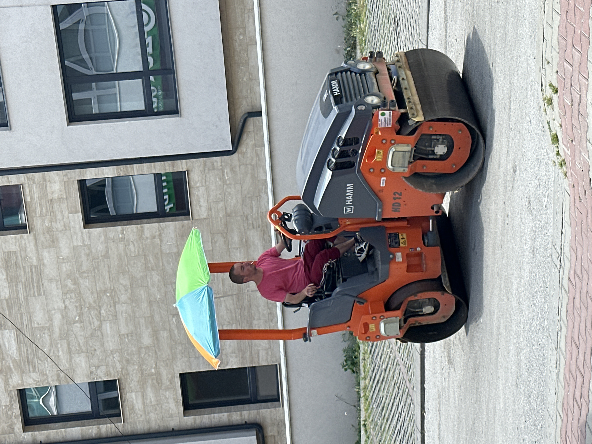 A man is operating an orange Hamm HD 12 roller on a paved surface. He is wearing a pink t-shirt and dark pants and has an umbrella attached to the roller for shade. The roller is in motion. In the background are modern buildings with stone and white facades and dark-framed windows. The scene appears to be in an urban setting, possibly a construction site or a newly paved street. The specific location in Veliko Tarnovo, Bulgaria, is not identifiable from the image alone.