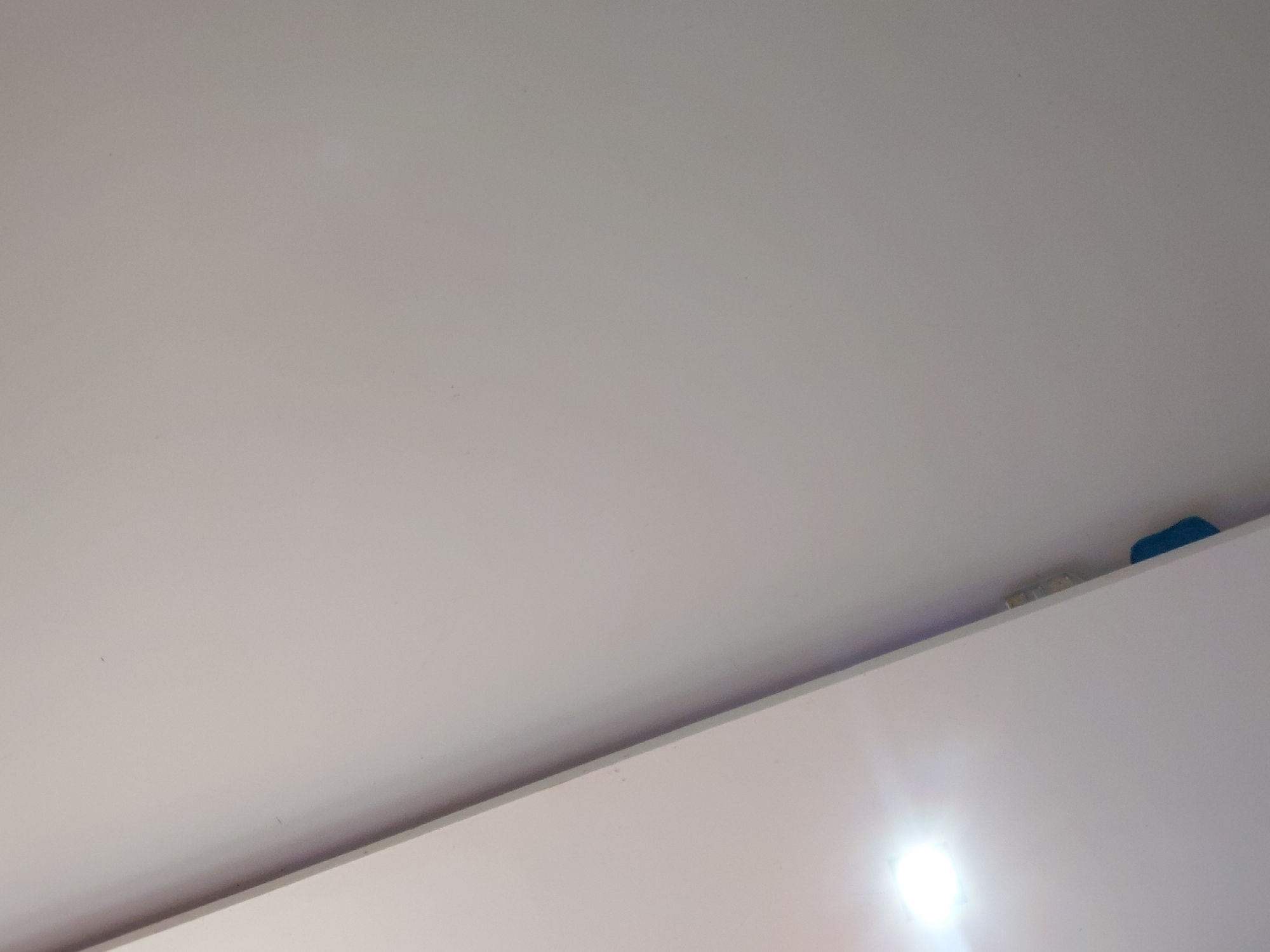 The image presents an upward-angled interior shot, primarily featuring a light-colored ceiling and the top edge of a white furniture piece or wall. The dominant upper and left sections of the frame are occupied by a smooth, off-white ceiling surface, appearing largely unadorned.
In the lower right corner, a powerful, bright light source is visible, casting a significant glare that illuminates the surrounding area. This light source appears to be part of an artificial lighting fixture. Below the ceiling and extending diagonally from the lower left to the right side of the image, there is a flat, white surface, which represents the upper section of an object, likely a cabinet, wardrobe, or a high shelf.
Along the narrow space or crevice where the ceiling meets the top of this furniture, two small objects are discernible towards the upper right corner. One is a distinct blue item, possibly a piece of fabric or a small plastic object, and next to it, a lighter, perhaps translucent or reflective object is tucked away.
No people or discernible activities are present. The scene is static and brightly lit by the artificial source, making the time of day unidentifiable. There is no visible text. The overall impression is of a clean, functional indoor space, located in Santa Ana, Venezuela.