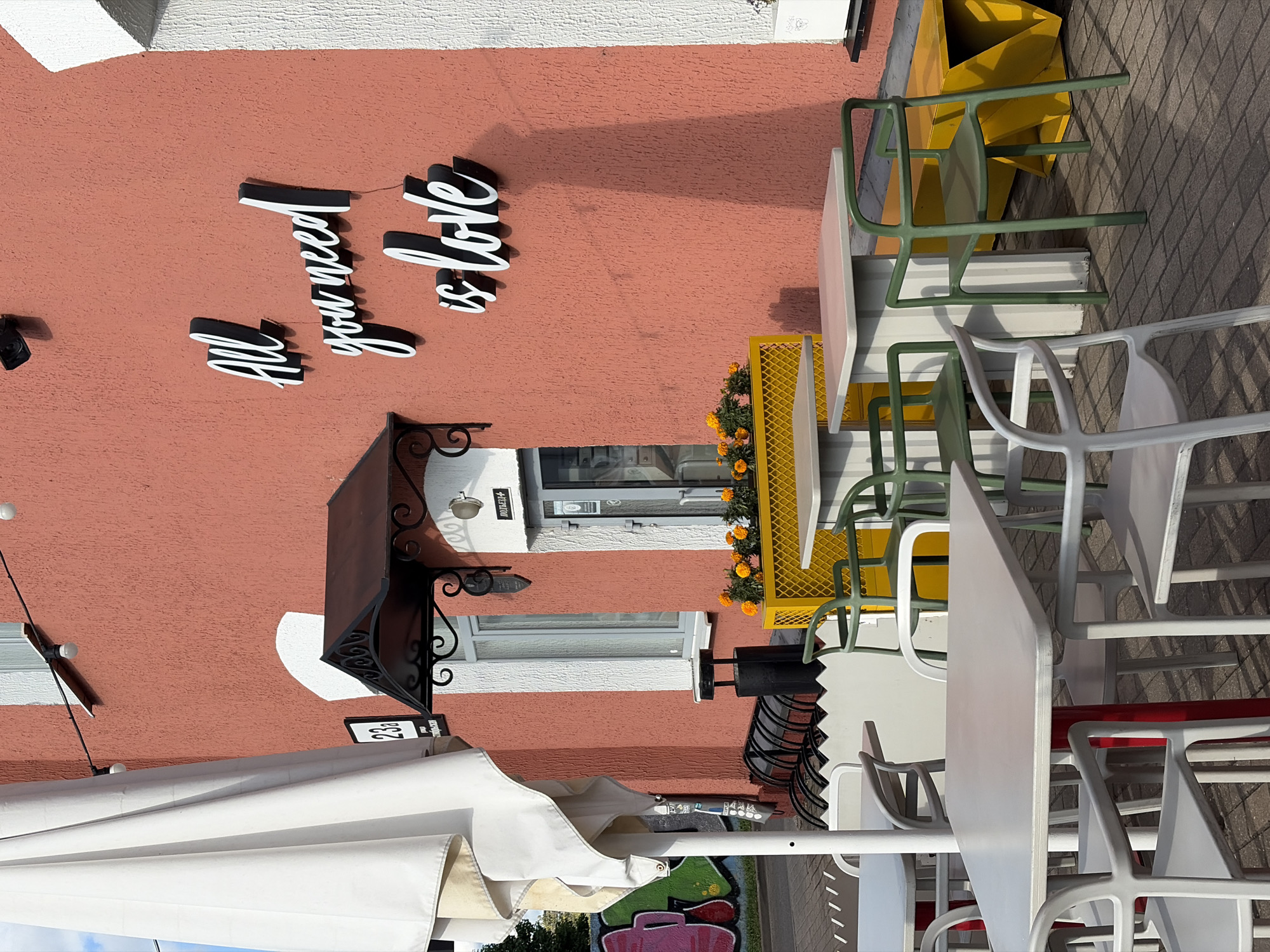 The image shows an outdoor seating area of a cafe or restaurant located at 23a Zybickaya Street in Minsk, Belarus. The building facade is a textured coral pink color. White three-dimensional lettering on the wall reads "All you need is love." In the foreground, several tables and chairs are arranged on a brick patio. The tables are light gray with red legs, and the chairs are a mix of light gray and dark green. Behind the tables, a yellow planter box with orange flowers is visible, along with a white awning over a doorway. To the left, a large white umbrella is partially visible. Graffiti art adorns a wall further in the background. The scene is brightly lit by sunlight, casting shadows across the patio. There are no people visible in the image.