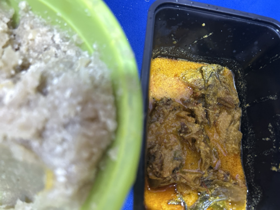 The image displays a top-down view of a meal in Ilishan, Nigeria. In the upper portion, a light green bowl contains a light beige, granular, semi-solid food substance, possibly a staple like pounded yam or fufu. Directly below it, a black rectangular container holds a stew or soup. This stew features a prominent reddish-orange liquid base and contains dark brown, fibrous solids, consistent with cooked meat or fish, along with visible green leafy components. The containers rest on a vibrant blue surface. No individuals or specific actions are depicted.