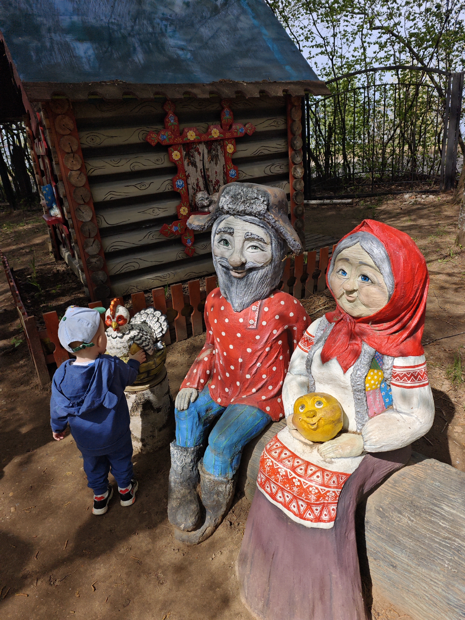 A full, medium-shot view of an outdoor display with a small child in front of it, taken in Chaikovsky, Russia. On the left stands a small wooden hut with detailed carvings around its front. A man statue sits to the right, wearing a red shirt with white polka dots, blue pants, and boots, with a grey hat, grey hair, and a grey beard. Beside him sits a woman statue with a red headscarf, holding a yellow orb with a face. A small boy in a blue outfit and a blue cap stands on the left, reaching towards a chicken statue. The ground is dirt with some fallen twigs and leaves. Trees and foliage are visible in the background behind the hut and statues.
