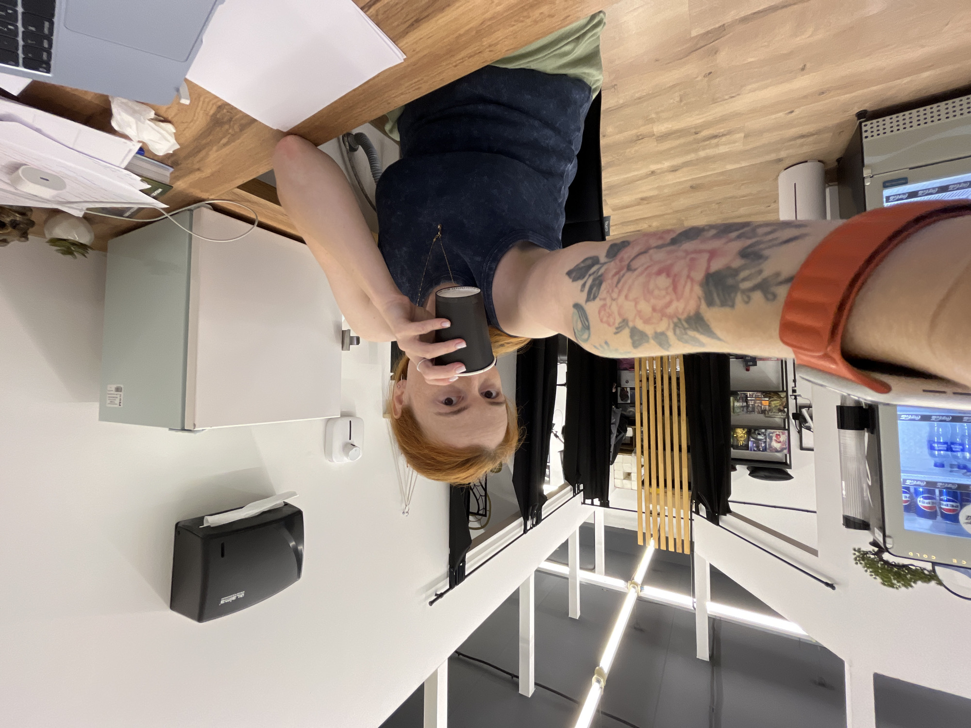 The image shows a woman sitting at a wooden desk, holding a cup. She has red hair and is wearing a dark blue tank top and green shorts. Her left arm, visible in the foreground, has a large tattoo of pink flowers and green leaves. The desk is covered with papers and a laptop. Behind her, a white mini-fridge and a black paper towel dispenser are mounted on the wall. To the left, there is a glass-fronted vending machine filled with drinks. The environment appears to be an indoor commercial space, possibly an office or retail store, with fluorescent lighting and wooden flooring. There is no indication of the location being in Chaikovsky, Russia.