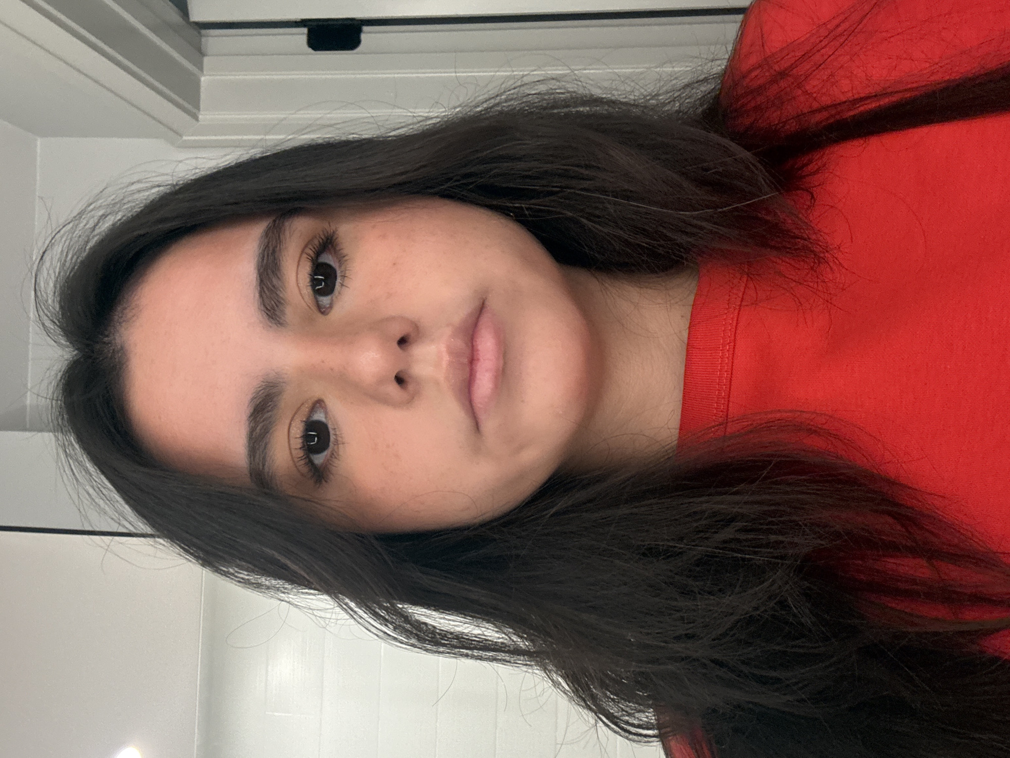 A close-up portrait shows a young woman with dark, shoulder-length hair and fair skin. She wears a red shirt. Her hair frames her face, and her eyes are dark. The background is a blurred white wall and a doorway with dark hardware. The location is not discernible from the image.
