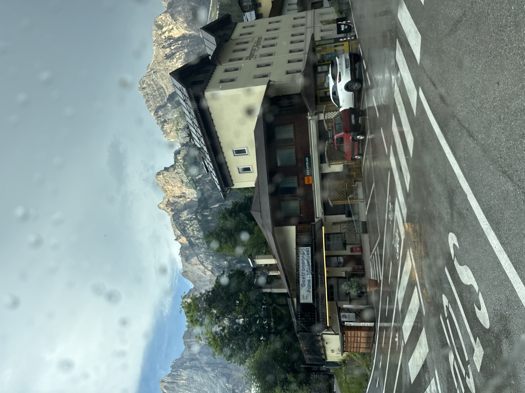 The image depicts a street scene in San Vito di Cadore, Italy, with a prominent view of the Dolomite mountains in the background. The foreground features a wet asphalt road with white crosswalk markings, indicating a pedestrian crossing. Two cars, a red compact car and a white SUV, are parked in a lot adjacent to a building. The building appears to be a multi-story establishment, possibly a hotel or apartment building, with a cream-colored facade and dark-pitched roofs. A sign above a ground-level storefront reads "Gastronomia Pane Minimarket." Another sign further down the building says "BAR ANTELAO." Green trees are visible between the buildings and the mountain backdrop. The sky is overcast with some patches of blue visible. The presence of water droplets on the camera lens suggests it may have been raining or the image was taken through a wet window.