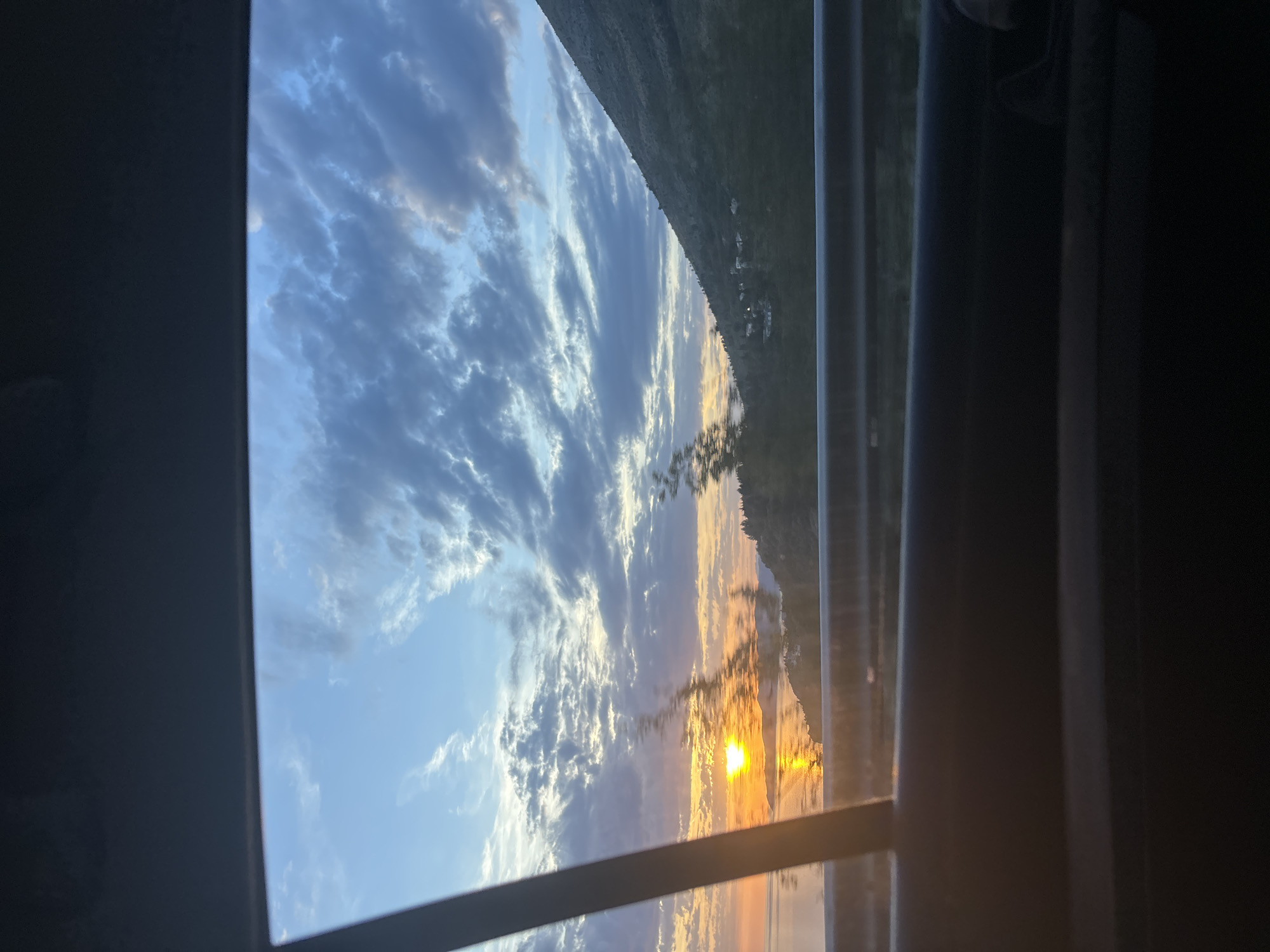 The image depicts a sunset over a body of water, likely the Adriatic Sea, observed from the interior of a vehicle on a coastal road near Petrovac na Moru, Montenegro. The sun, a prominent yellow orb, is situated on the left, casting an intense, reflective path of orange and yellow light across the water surface. The sky exhibits a varied cloud structure, with lighter, wispy clouds against a blue sky in the upper left transitioning to denser, darker, and more stratified cloud formations occupying the majority of the upper frame, illuminated by the low sun. A large, dark, and densely vegetated mountain slope rises abruptly on the right side of the frame, extending from the lower mid-section to the top right corner. Scattered small, light-colored buildings are discernible on the lower flanks of this mountain. In the immediate foreground, dark elements of the vehicle's interior are visible: a vertical pillar on the far left, a horizontal guardrail (likely roadside or bridge railing) running across the lower third of the image, and a dark car door panel or dashboard along the bottom. Blurred foliage is present in the mid-ground, indicative of movement. No discernible human figures are present.