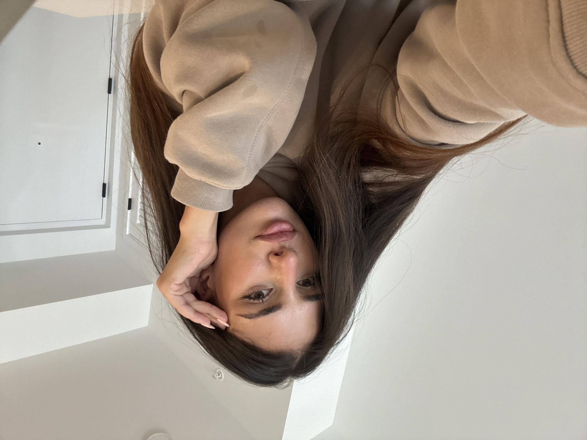 A young woman with long, dark brown hair and light skin is pictured in a close-up, waist-up shot. She is wearing a tan hoodie. Her right hand is raised to her face, with her fingers resting on her temple. She is looking directly at the camera with a neutral expression. The background appears to be an indoor setting, with a plain white wall and a white door visible. The lighting is bright and diffused. The image is taken from a slightly high angle.