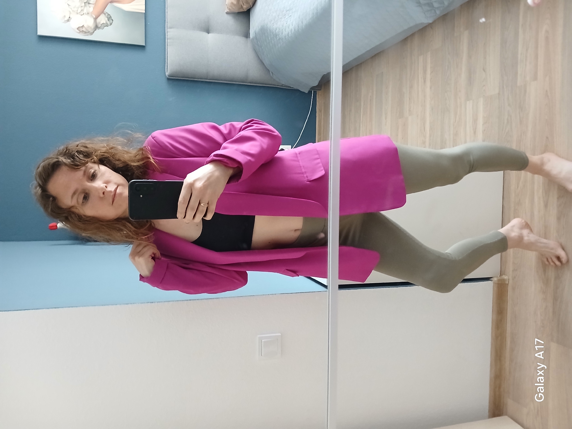A woman is taking a selfie in a mirror. She is wearing a pink blazer, a black top, and olive-colored leggings. She is holding a black smartphone in her hand. The background includes a blue wall with artwork, a gray couch, a light switch, and a wooden floor. "Galaxy A17" is visible on the bottom right of the image. The location is likely a domestic interior rather than a specific outdoor location in Yekaterinburg, Russia.