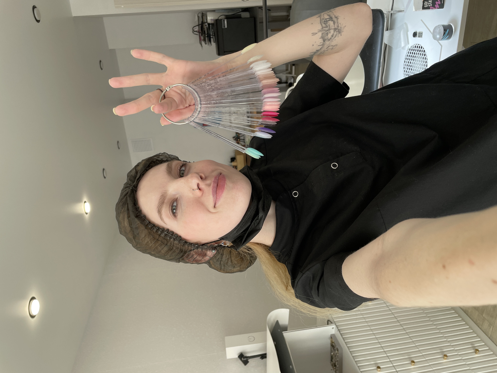 A fair-skinned person with blonde hair is in what appears to be a salon setting. They are wearing a dark protective hair covering and a black garment with buttons, and a face mask is partially visible. The person is holding up a color palette of nail polish samples with their left hand, making a peace sign with their fingers. There is a tattoo on their right arm. The background includes salon equipment, such as a chair and possibly a nail station, under what appears to be fluorescent overhead lights. There is nothing indicating that the location is in the city of Izhevsk, Russia.