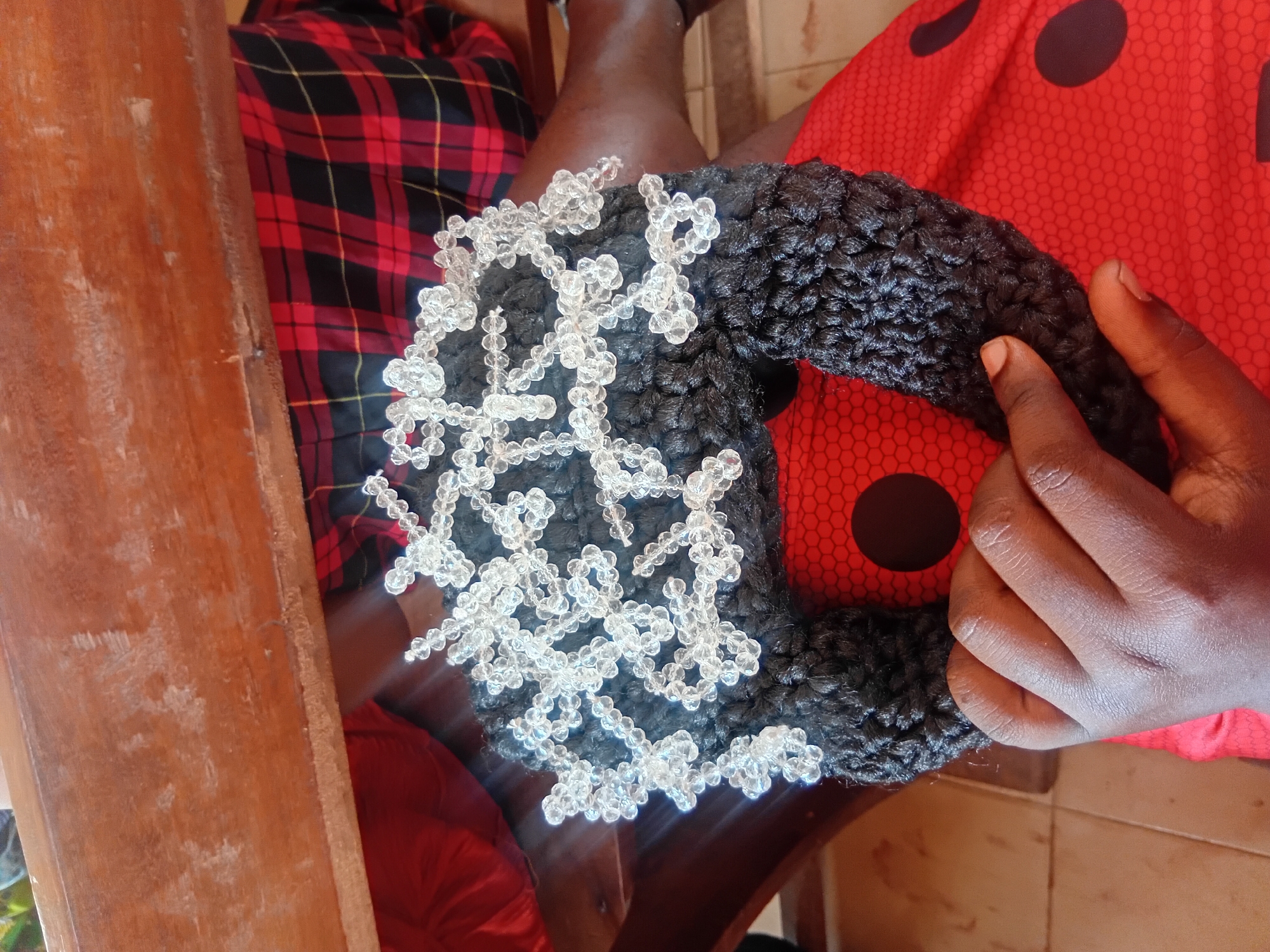 A person's hands are holding a handmade object. The object is black and appears to be crocheted, with clear beads attached to it in a decorative pattern. The person is wearing a red top with black polka dots. In the background, a person is wearing a red and black plaid shirt and sitting on a wooden bench. The scene appears to be indoors or in a shaded outdoor area, with a wooden surface visible above the subjects. The floor is tiled. The image was taken in Ejule, Nigeria.