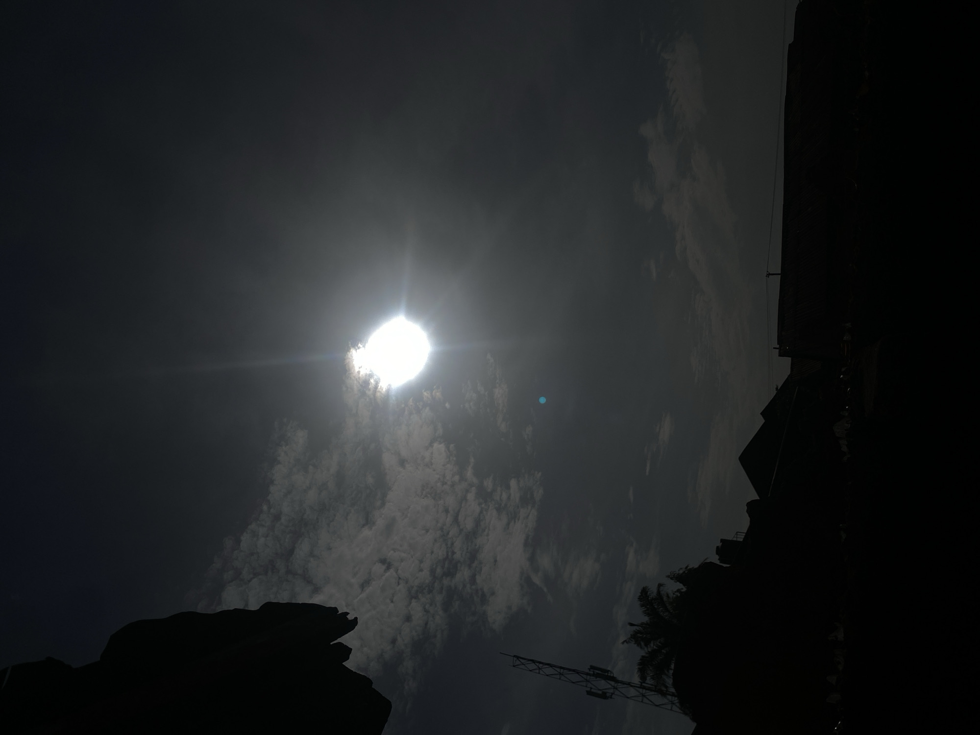 The media file displays a dramatic, high-contrast scene dominated by the sky and silhouetted structures, captured in a portrait orientation that appears to be rotated clockwise by 90 degrees from a typical landscape view.
In the upper-left portion of the frame, a brilliant, overexposed sun shines intensely through a layer of white and grey clouds, creating a powerful lens flare that extends horizontally from its core. The clouds around the sun are thick and textured, transitioning to darker, more diffused cloud formations and a deep grey-blue sky towards the top and center-right. A small, faint blue-green speck, possibly another lens artifact, is visible to the upper right of the sun.
The lower-right and bottom edges of the image are occupied by stark, dark silhouettes of various structures. These include the jagged outlines of multiple buildings, some with discernible rooflines, that extend vertically along the right side and horizontally across the bottom. Prominently featured in the lower-right is the distinct silhouette of a palm tree, its fronds sharply defined against the sky. Near the palm tree and extending further left along the bottom, a skeletal metal structure, possibly a communication tower or utility mast, is also silhouetted.
The intense backlighting from the sun renders all foreground elements as deep black silhouettes, creating a striking contrast with the bright sky and clouds. The time of day appears to be during daylight hours, likely midday or early afternoon, given the sun's brightness and position. The weather is partly cloudy to overcast, with the sun breaking through. No discernible human activity or visible text is present. The scene captures an atmospheric view of Barden Yana, Nigeria, with its characteristic structures and natural elements under a bright, cloud-filtered sun.