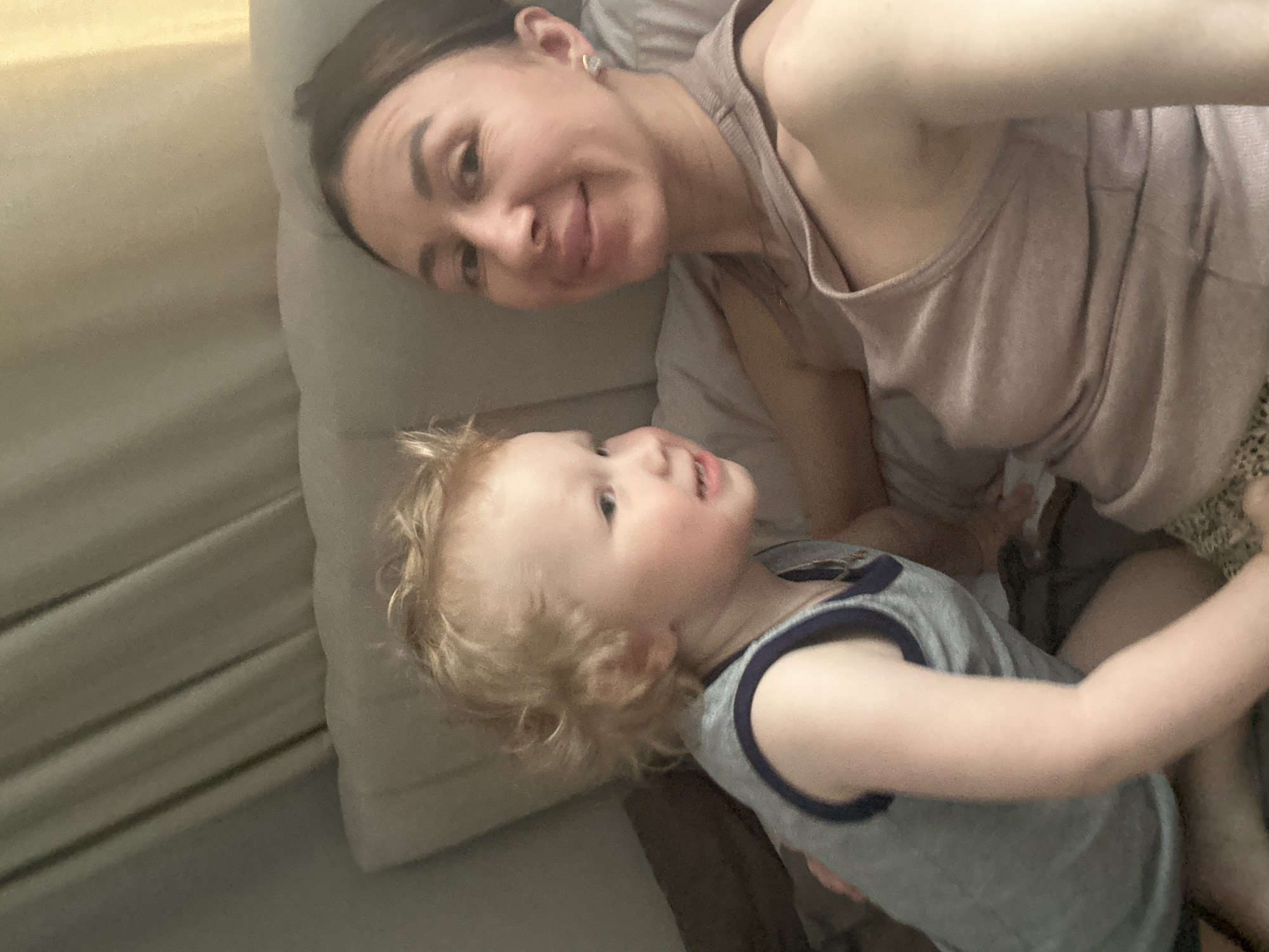 A woman and a young child are lying on a bed. The woman, with fair skin and dark hair pulled back, is wearing a light-colored tank top. She is smiling and looking at the camera. The child, who has light skin and curly blonde hair, is wearing a grey tank top with blue trim. The child is looking up at the woman. The background consists of a light-colored pillow and bedding. It is not possible to determine the location of this image as being in Votkinsk, Russia or the environment, using the content of the image.