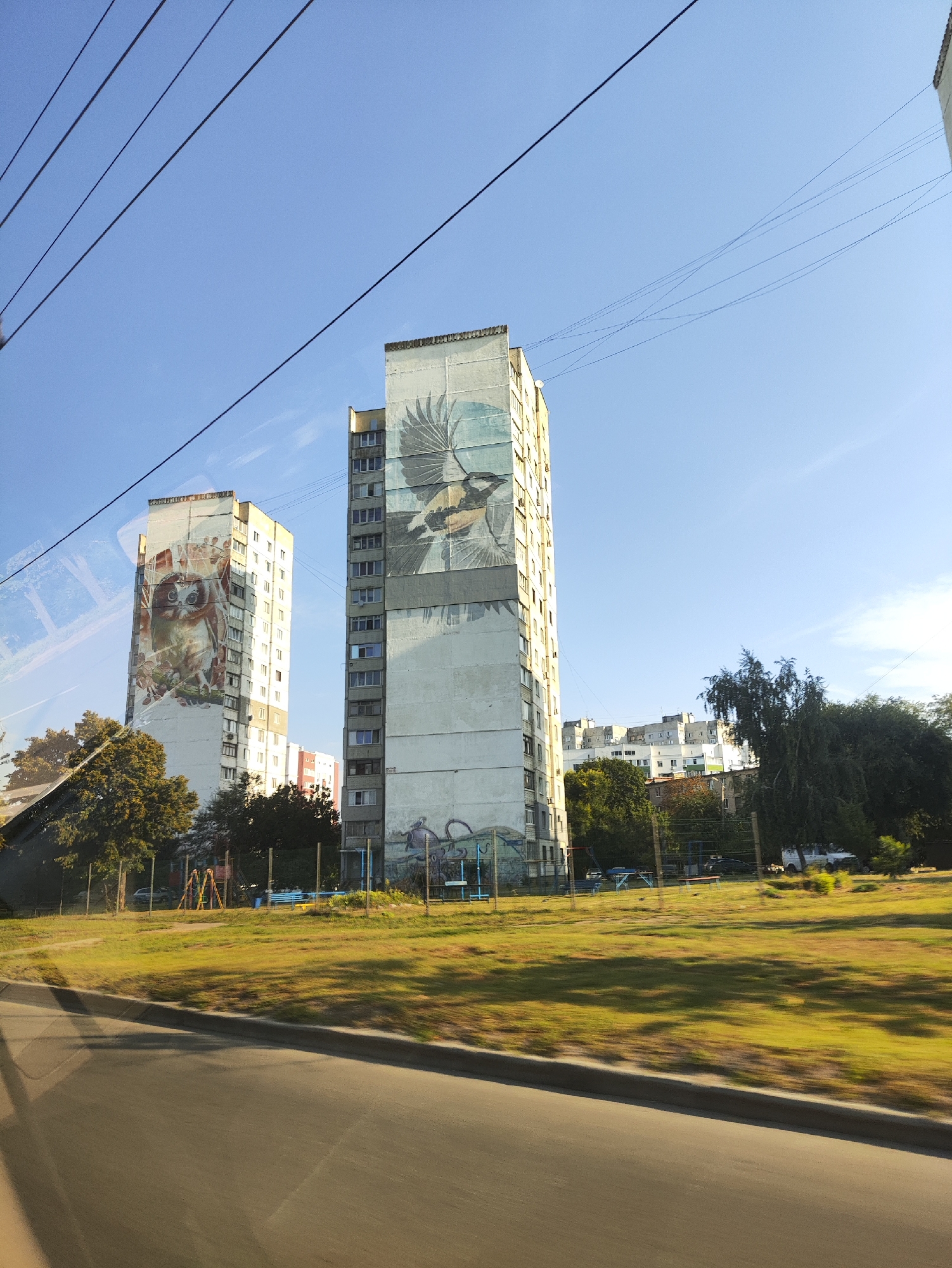 The image shows two tall, multi-story buildings in a city, with one building featuring a large mural of an owl and the other a mural of a bird in flight. The buildings are located in Kharkiv, Ukraine, specifically in the Saltivskyi district. The foreground shows a road with a blurred motion effect, indicating the photo was taken from a moving vehicle. Behind the buildings, there are other residential buildings and lush green trees. A playground with swings and a slide is visible in the middle ground, along with a fenced-off grassy area. The sky is clear and blue, suggesting a sunny day. The architecture of the buildings suggests a post-Soviet era construction.
