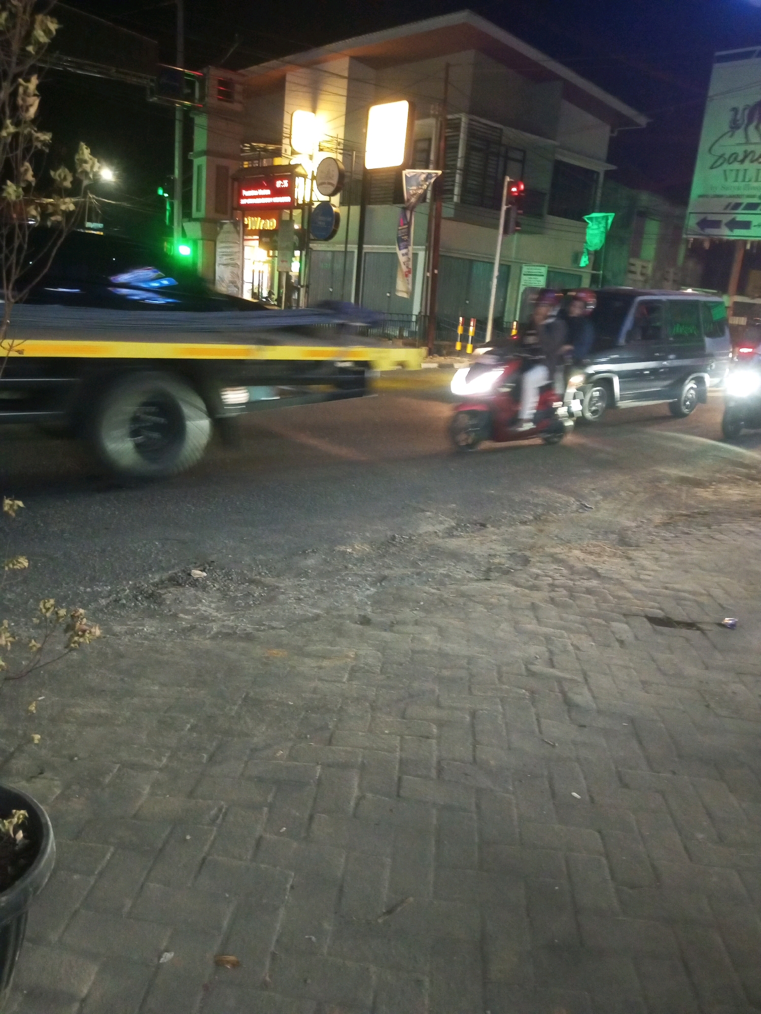 The image depicts a nighttime street scene in Mojotengah, Indonesia, with several vehicles and a building. In the foreground, a tow truck with a yellow stripe is parked on the right side of the road. A red scooter with two occupants, one driving and one passenger, is moving in front of the tow truck. Behind the scooter is a dark-colored SUV. The building in the background has multiple stories with illuminated windows and signage. One sign, partially visible, appears to be for "Sans Village". The road surface is paved with interlocking bricks. A traffic light is visible, showing a red signal. The scene suggests a busy urban environment with ongoing traffic.