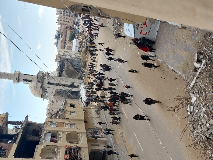 This image captures a severely damaged urban street in Khan Yunis, Palestinian Territory, viewed from an elevated perspective. In the background, a large mosque, identifiable by its prominent minaret, stands significantly damaged, with large sections of its structure collapsed and the dome scarred. To the left, a multi-story residential building exhibits extensive destruction, including exposed interiors, broken balconies, and visible burn marks. The street is an asphalt road, partially covered with dust and light debris, with faded white lane markings.
Hundreds of individuals, consisting of adults and children in civilian clothing, traverse the street, walking in various directions. In the mid-ground, adjacent to the damaged mosque, several white tent-like structures are visible amidst piles of rubble. Overhead power lines crisscross the sky. The immediate foreground shows substantial piles of concrete, rebar, and construction debris, indicating a collapsed or damaged structure below the viewpoint. A portion of another damaged building facade, featuring a white sign with red Arabic text, is visible on the right side of the image. The sky above is partly cloudy.