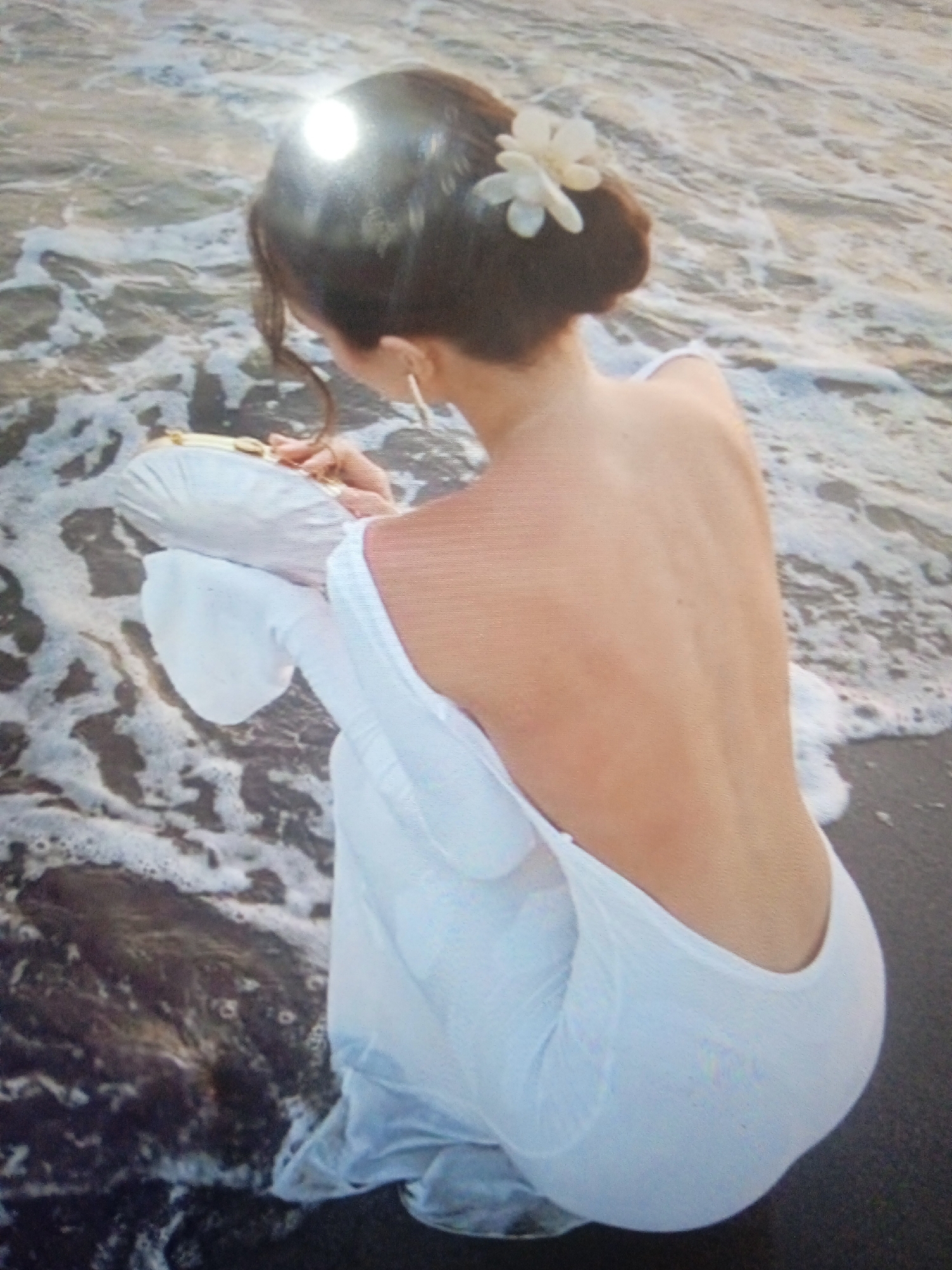 A woman wearing a white dress is sitting by the sea. She has a flower in her hair and is holding a white clutch bag. The water is lapping at her feet. The image is taken from behind the woman, showing her bare back and the waves of the sea. The scene is peaceful and serene, with the sound of the waves and the gentle breeze. The location is not specified in the city of Chernivtsi, Ukraine.