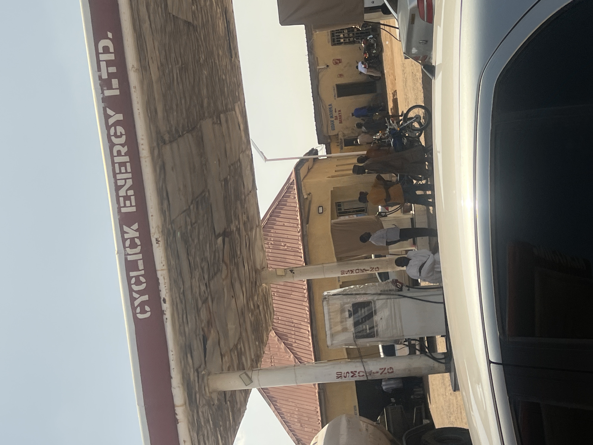 This image depicts an outdoor scene at the Cyclick Energy Ltd. gas station in Gombe, Nigeria, under bright daylight. The foreground is partially occupied by the roof and a dark window of a light-colored sedan. Above, a gas station canopy, visibly worn with peeling material on its underside, displays the name "CYCLICK ENERGY LTD." in white stencil lettering against a reddish-brown band. Two white support pillars for the canopy are visible, each bearing faded red vertical text "NO SMOKING."
In the middle ground, multiple individuals, predominantly males dressed in various attire including traditional garments, are gathered near and between gas pumps. One gas pump on the left appears wrapped in plastic or sheeting, while another to its right is uncovered and operational. A large white tanker truck is partially visible on the far left. Several motorcycles are parked or positioned around the people. One man in a white garment is crouched near a pump.
In the background, two single-story buildings are visible. The closest has a reddish-brown corrugated roof and yellow walls. Further back and to the right, another yellow-walled building with a darker, flatter roof displays text including "UDAN MANNA DA BAHAYA" and "FEMALE TOILET" with an arrow. The ground across the scene is dry and dusty.