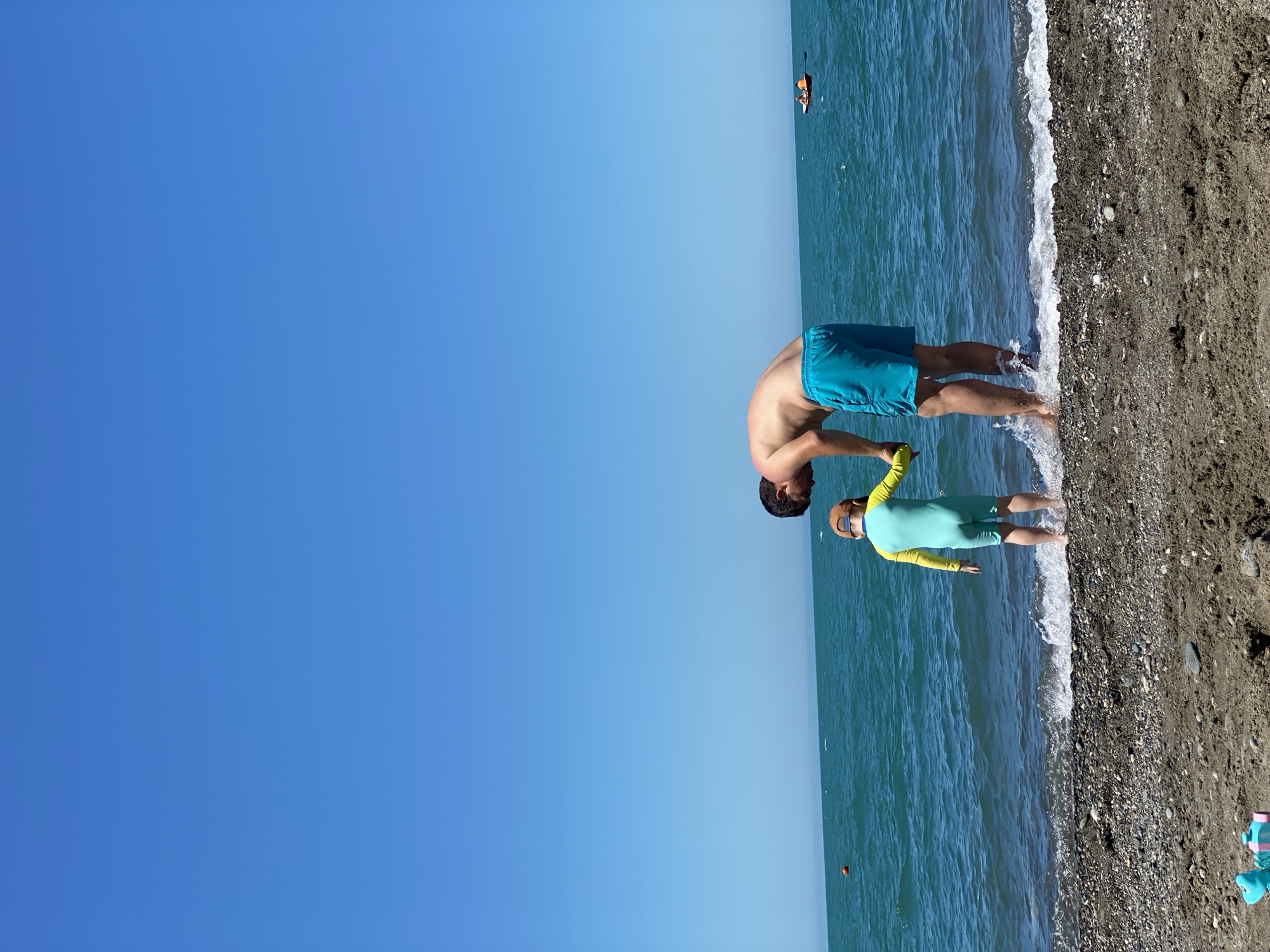 The image depicts a beach scene in Sochi, Russia, on a clear, sunny day. An adult male and a young child are standing at the water's edge, viewed from behind. The man, shirtless and wearing light blue swimming shorts, is bent forward, holding the child's right hand. The child is clad in a full-body, long-sleeved swimsuit, light blue on the lower body and lime green/yellow on the upper body and sleeves, with a brown baseball cap worn backward. Small waves are breaking around their feet on the dark, coarse sand and pebble beach. The sea is a vibrant turquoise-blue. In the mid-distance to the left, an orange spherical buoy is visible on the water. To the far right, a small white boat with two individuals aboard is present on the horizon. The sky is a uniform, clear bright blue, occupying the upper two-thirds of the frame. A light blue and pink plastic beach toy is partially visible on the sand in the bottom-left corner.