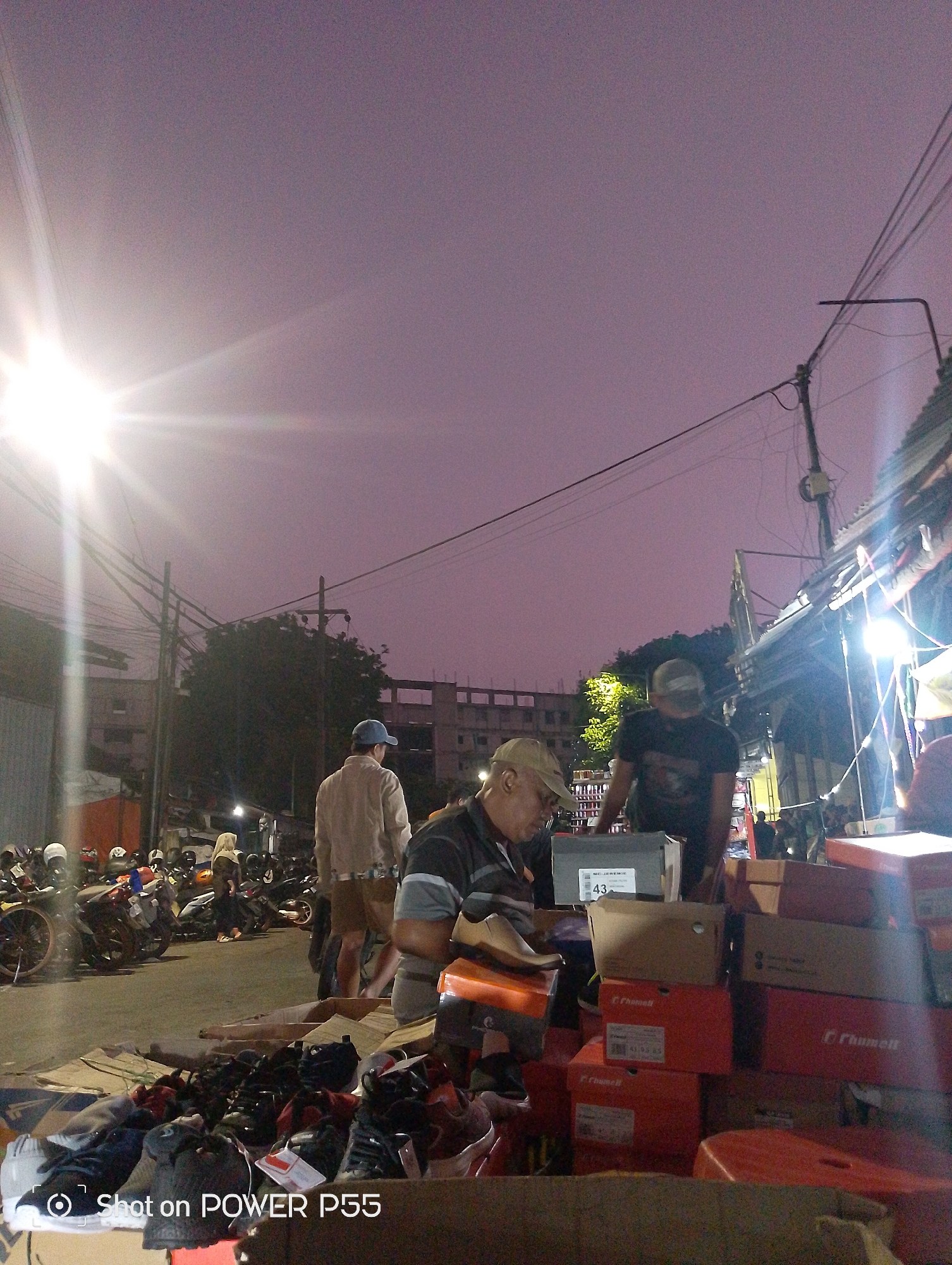 The image depicts an outdoor market scene at dusk in Surabaya, Indonesia. The sky is a deep purple, indicating the transition from day to night, and a bright street light casts a strong glare on the left side of the frame. Several people are present, some appearing to be vendors and others customers, engaged in activities related to commerce. In the foreground, a large assortment of footwear, including shoes and sneakers, are displayed on cardboard and on the ground, suggesting a shoe market. Several stacked cardboard boxes, some labeled with branding such as "Rhumell", are also visible, likely containing more merchandise. In the background, a street lined with parked motorcycles suggests this is a busy thoroughfare that has been converted for market use. Buildings and utility poles are visible, common elements of an urban environment. The overall impression is one of a bustling night market in Surabaya.
