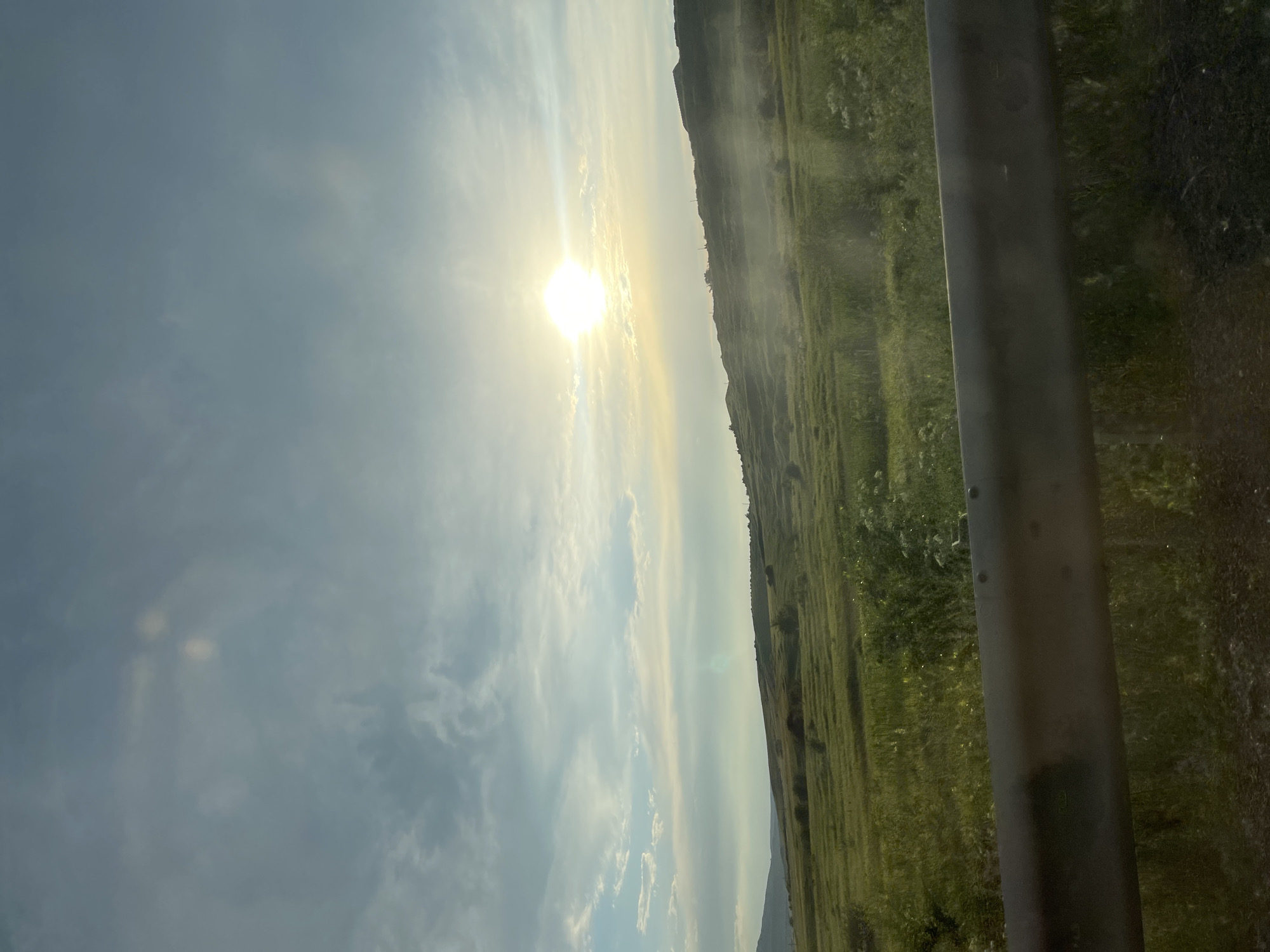 The image shows a landscape with a bright sun in a cloudy sky. The sun is positioned on the left side of the frame, casting rays of light. A hillside covered in green grass slopes downwards from the right side of the image. There is a dark, vertical object on the far right, which appears to be a pole or a fence. The overall scene suggests an outdoor setting, possibly a rural area, with a sense of natural light. The time of day appears to be either sunrise or sunset due to the low angle of the sun and the warm light. The weather is partly cloudy. The location is identified as Tatarka, Russia. There is no discernible activity or event taking place, and no visible text.