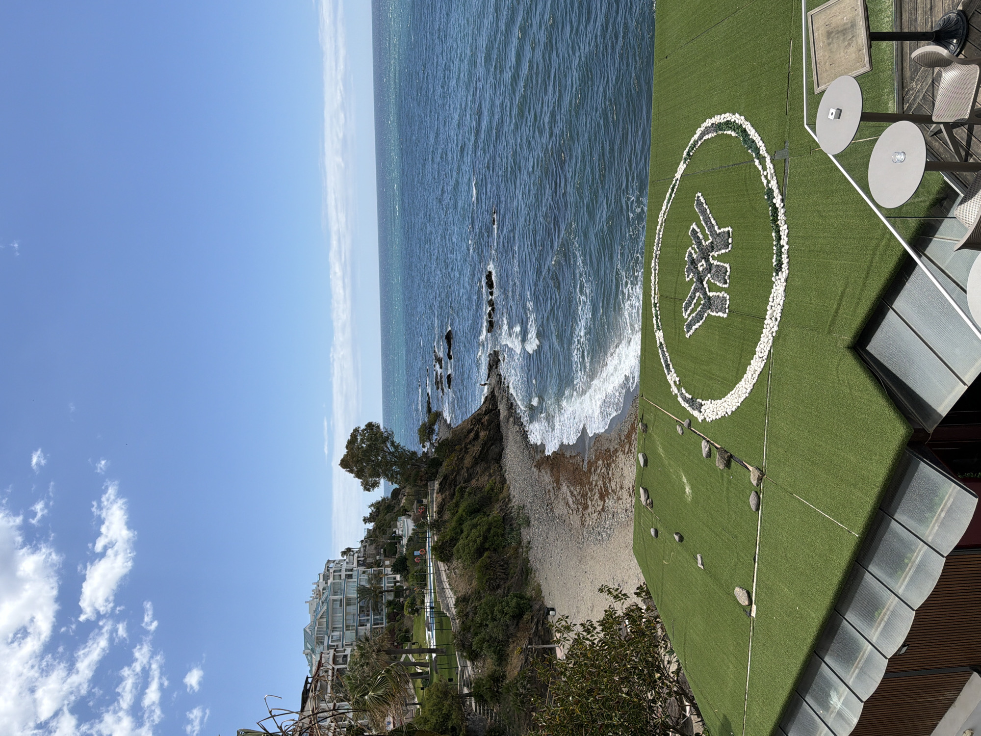 The image captures a view of the coastline in Benalmádena, Spain, under a partly cloudy sky. The scene encompasses the ocean, a beach, buildings, and a helipad on a rooftop. The ocean has visible waves breaking near the shore, which is a mix of sand and rocks. The beach is narrow, leading to a cliff or hillside with vegetation. On the left side of the frame, there are residential buildings. A rooftop helipad with a design made of white rocks is on the right. The helipad is surrounded by artificial grass and has some scattered stones. There is also an outdoor seating area with tables and chairs.