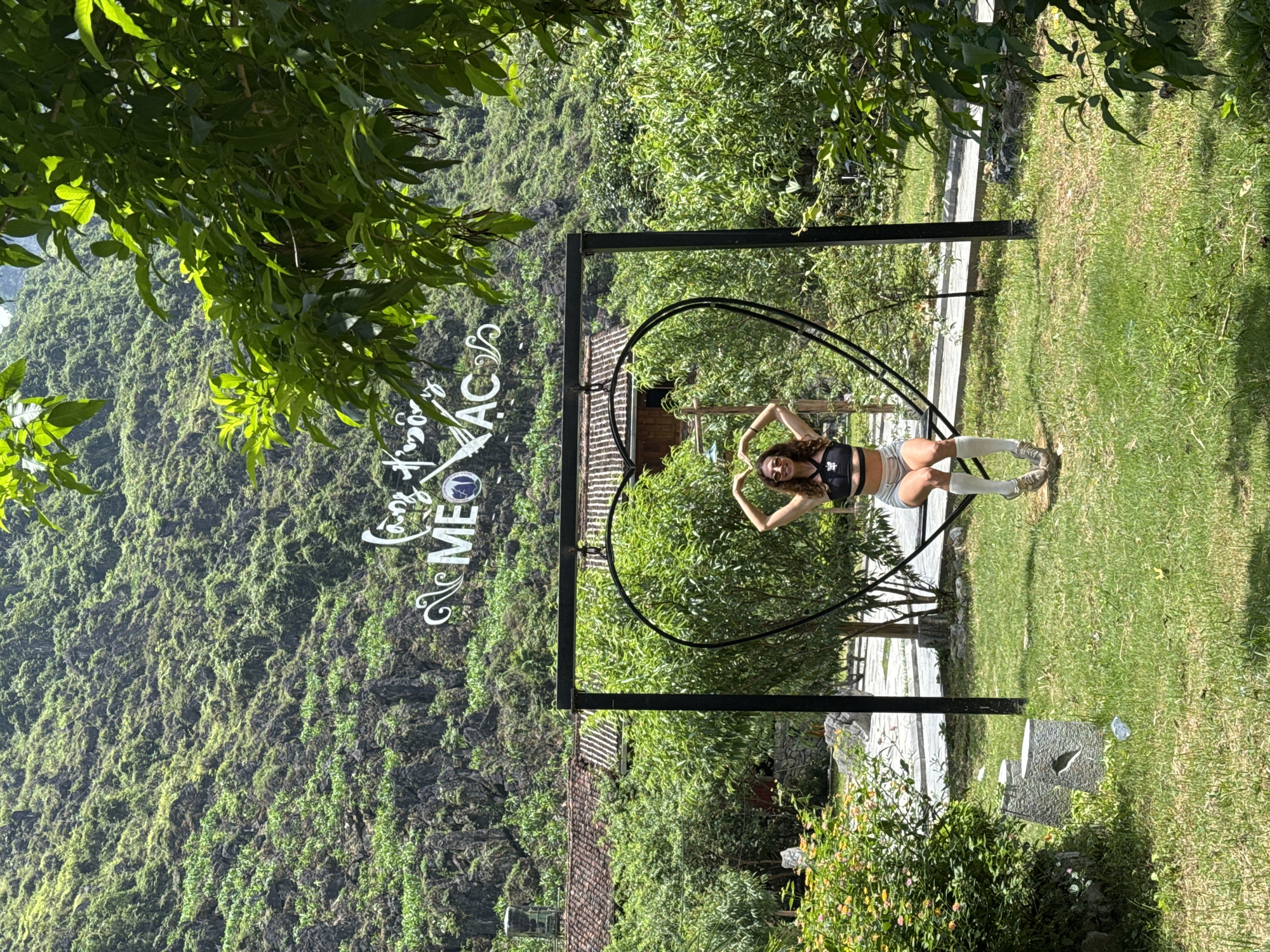 The image shows a woman sitting in a heart-shaped swing in Mèo Vạc Commune, Vietnam. She is wearing a black top, grey shorts, and long socks. She has her arms raised above her head. Behind her is a lush, green hillside. There is a sign that reads "Mèo Vạc" in Vietnamese. The swing is made of black metal and is positioned on a grassy lawn. There is a building with a red tiled roof in the background.