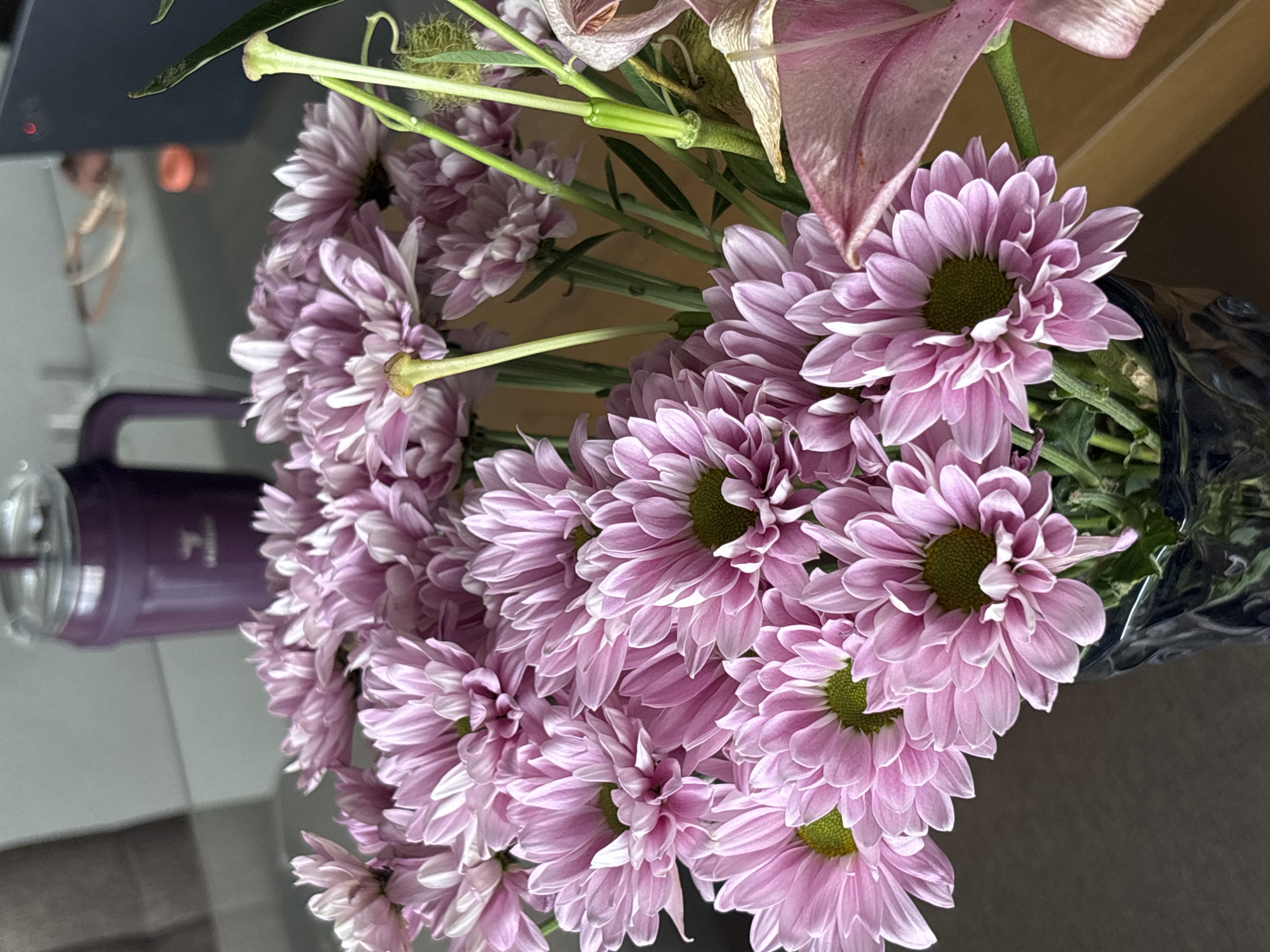 A bouquet of purple chrysanthemum flowers and a faded pink lily are arranged in a dark-colored vase on a table. A purple and clear water dispenser is visible in the background, situated on a glass table. The scene is indoors and the precise location within Mexico City, Mexico, cannot be determined from the image. No people are visible. The primary objects are the flowers, vase, and water dispenser. There are no specific actions or interactions depicted.