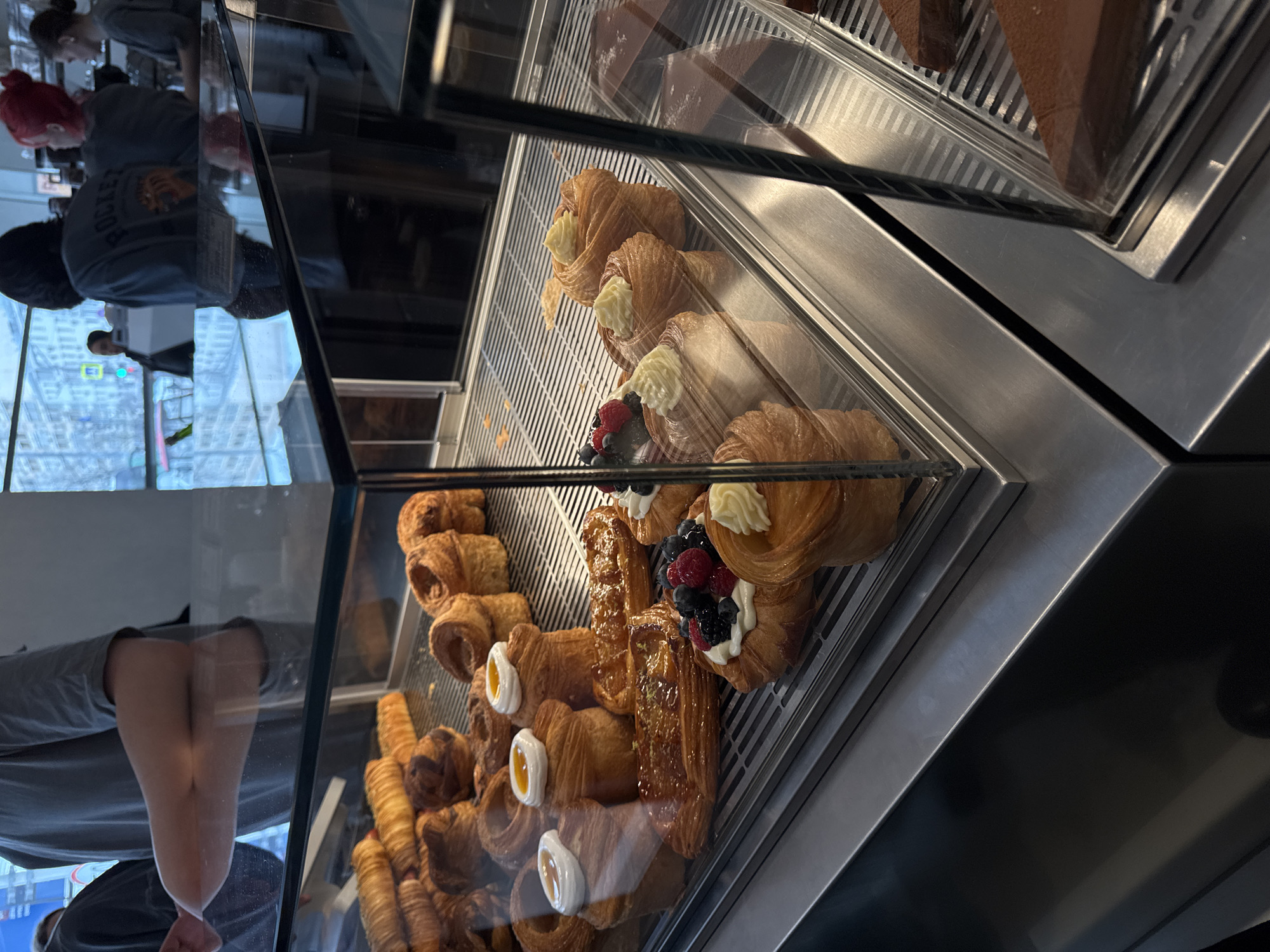 The image shows a refrigerated display case filled with an assortment of pastries. The pastries include croissants topped with cream and various fruit. Some croissants are filled with custard or cream. In the upper section, there is chocolate cake. Reflections are visible in the glass display. Reflected in the glass are people near a window. Given the architectural style and window view, this image could be located in a shop at the top of the Federation Tower in the Moscow City business district of Moscow, Russia.