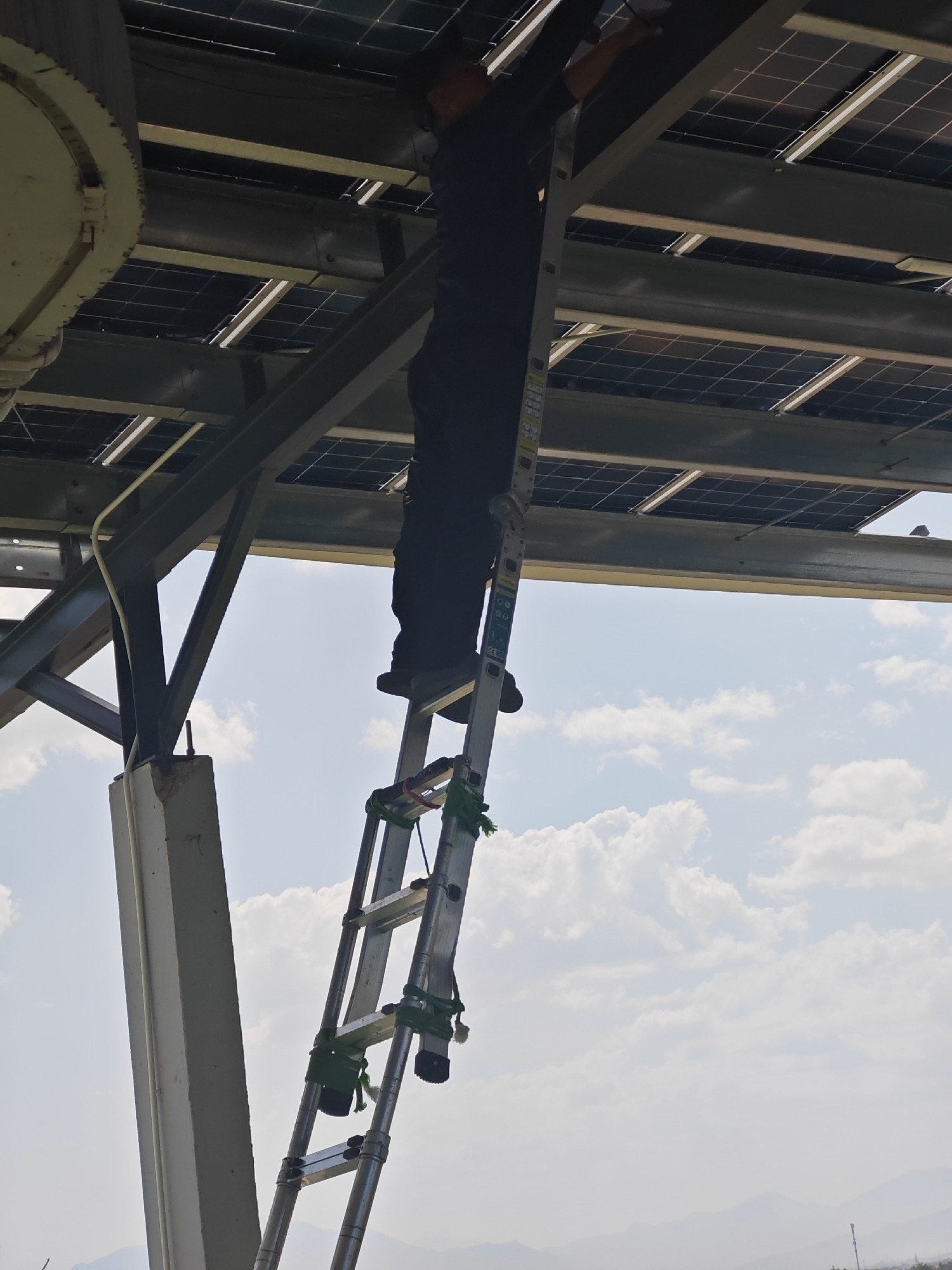 A person in dark clothing is on a ladder, working on solar panels. The solar panels are mounted on a metal structure above the person. The sky is visible in the background, with some clouds. The image appears to have been taken outdoors, likely during the day. The presence of solar panels suggests a location that utilizes renewable energy. The context of the image, with a person working on the panels, indicates maintenance or installation activity.