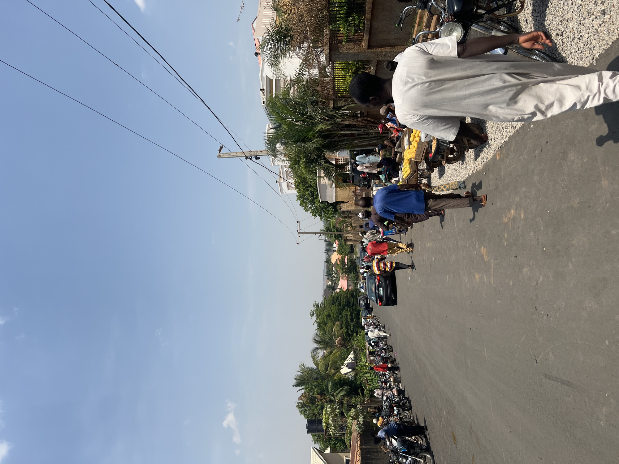 A street in Abuja, Nigeria shows a line of motorcycles parked along the right side of the road. Several people are walking along the road, and some appear to be interacting with a vendor. A black car is visible further down the road. Palm trees and other vegetation are on both sides of the street, with buildings in the background. Overhead power lines stretch across the sky, which is blue with few clouds.