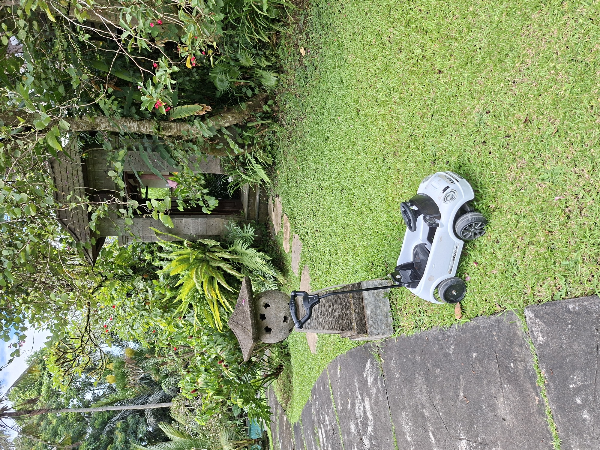 The image depicts a backyard scene in the Ubud District of Indonesia. Lush greenery, including trees, ferns, and other plants, surrounds a small stone gazebo-like structure. A gray stone lantern stands near the gazebo. A white toy car is parked on the grass next to a stone pathway. The overall environment suggests a tropical or subtropical climate. There are no people visible in the scene.