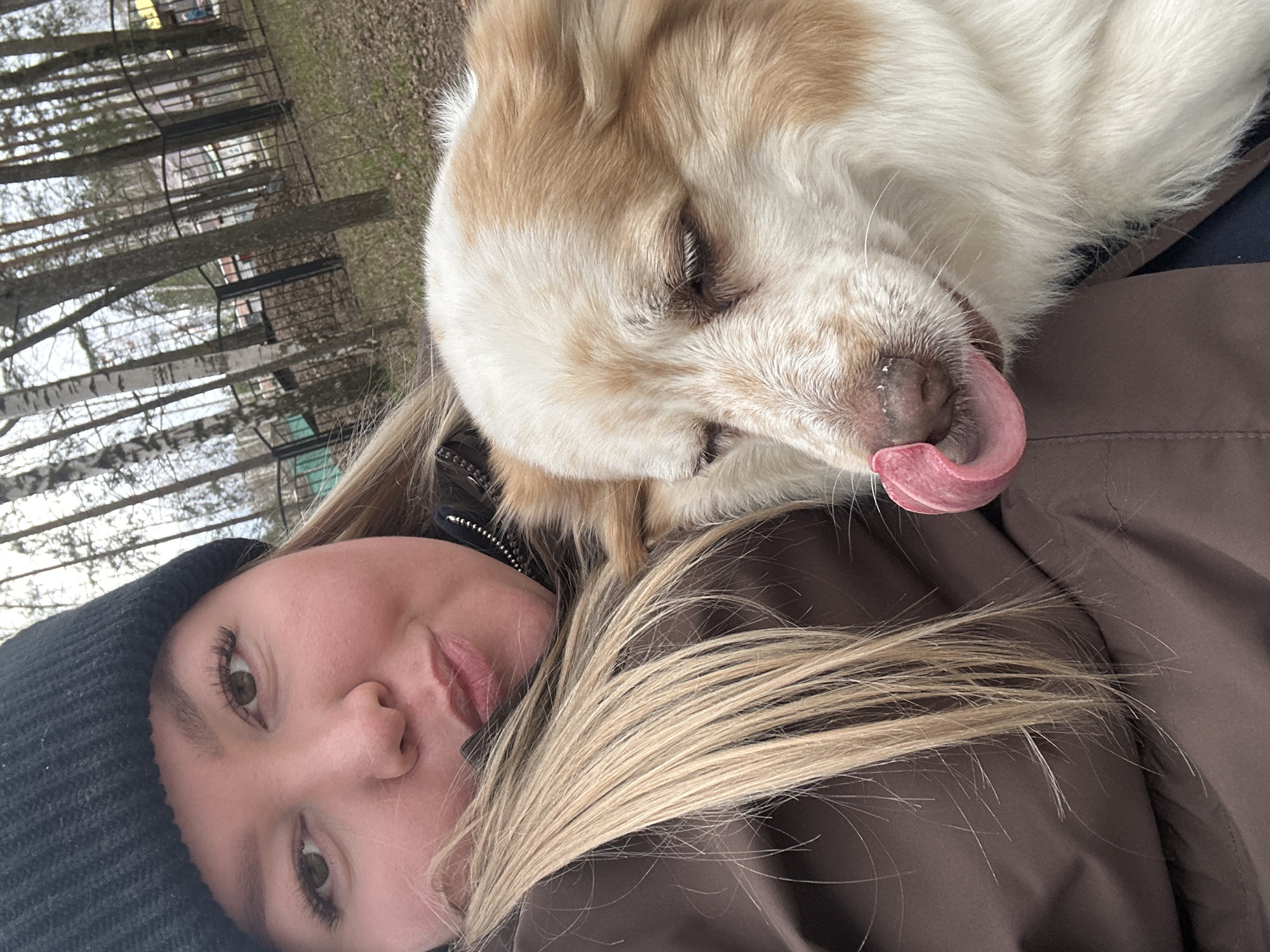 This close-up, rotated image captures a serene outdoor moment between a woman and her dog in Chaikovsky, Russia. The woman, lying horizontally, has fair skin, light-colored hair, and a neutral expression with slightly parted lips. She wears a dark knitted beanie and a dark brown or black jacket, suggesting cool weather.
Cuddled close to her, with its head resting on her shoulder or chest, is a fluffy dog with prominent white and light brown fur. The dog's pink tongue is extended, appearing to lick its nose or just hanging out casually, conveying a sense of contentment or playfulness. Its fur appears thick and soft.
The background is a soft blur of an outdoor environment, dominated by the bare trunks and branches of numerous trees, indicating late autumn, winter, or early spring. Faint structures or fences are visible through the trees in the distance. The natural lighting suggests it is daytime, possibly under an overcast sky. No visible text is present in the image. The scene portrays a tender and relaxed connection between the human and animal subjects in a quiet, natural setting.