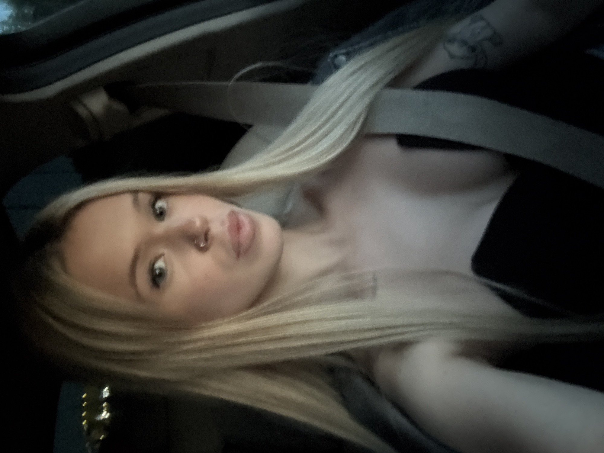 The image is a close-up, vertically oriented photograph of a young woman with blonde hair, taken in what appears to be the interior of a vehicle. She is wearing a black top and a grey seatbelt is visible across her chest. Her hair is long and blonde, framing her face and extending across the frame. She has a nose ring and her lips are slightly parted. Her eyes are open and she is looking towards the camera, with a neutral expression. There is a tattoo visible on her upper arm. The lighting suggests it may be daytime, but the overall mood is somewhat dark and intimate. The background is indistinct, but the presence of the seatbelt and car interior strongly indicates she is a passenger in a moving vehicle. The scene is likely casual, perhaps a selfie taken during a car ride. There is no visible text in the image.