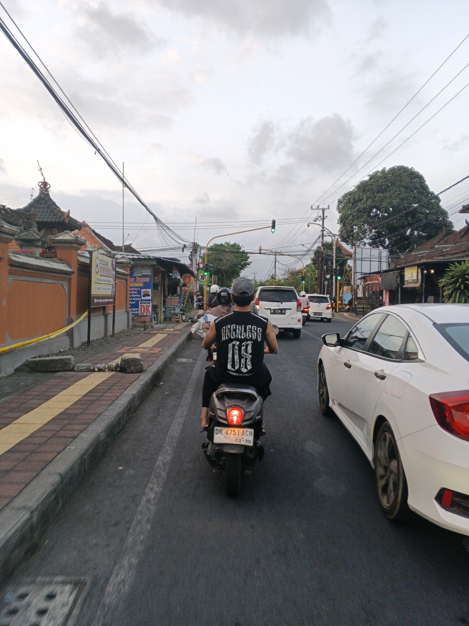 The image captures a street scene in Denpasar, Indonesia, during what appears to be a traffic jam. In the foreground, a person wearing a black t-shirt with "RECKLESS 08" printed on it is riding a scooter. The scooter's license plate reads "DK 4751 ACH". To the right of the scooter, a white car is visible, its rear portion dominating the right side of the frame. In the background, there are other vehicles, including white cars and scooters, stopped in traffic. The street is lined with buildings, shops, and an orange wall with ornate architectural details. Overhead, numerous power lines crisscross the sky, which is overcast. Traffic lights are visible, with one showing green. The overall impression is one of a busy urban street with moderate traffic congestion.