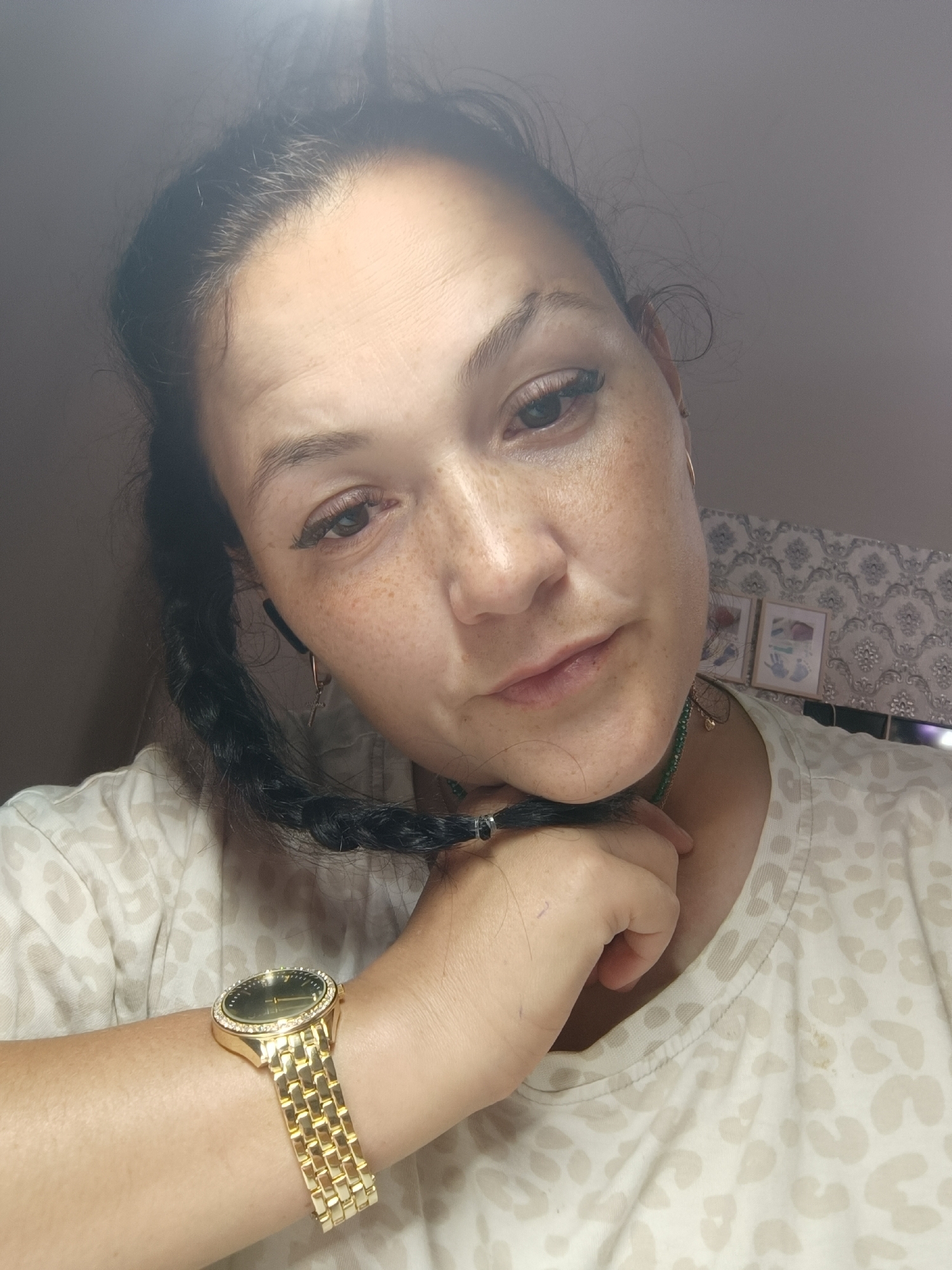 A woman is shown in a close-up, selfie-style shot. She has brown hair pulled back and a braid that extends across her chest. She is wearing a light-colored t-shirt with a subtle animal print pattern and a gold watch on her left wrist. Her right hand is raised and touching her neck, with the braid wrapped around her fingers. The background is blurred, but appears to be an indoor setting with a patterned wallpaper and two framed pictures visible on the wall. The image is lit from above, creating a bright, hazy effect in the upper portion of the frame. The overall impression is casual and personal. The location in Chaikovsky, Russia is not evident from the image.