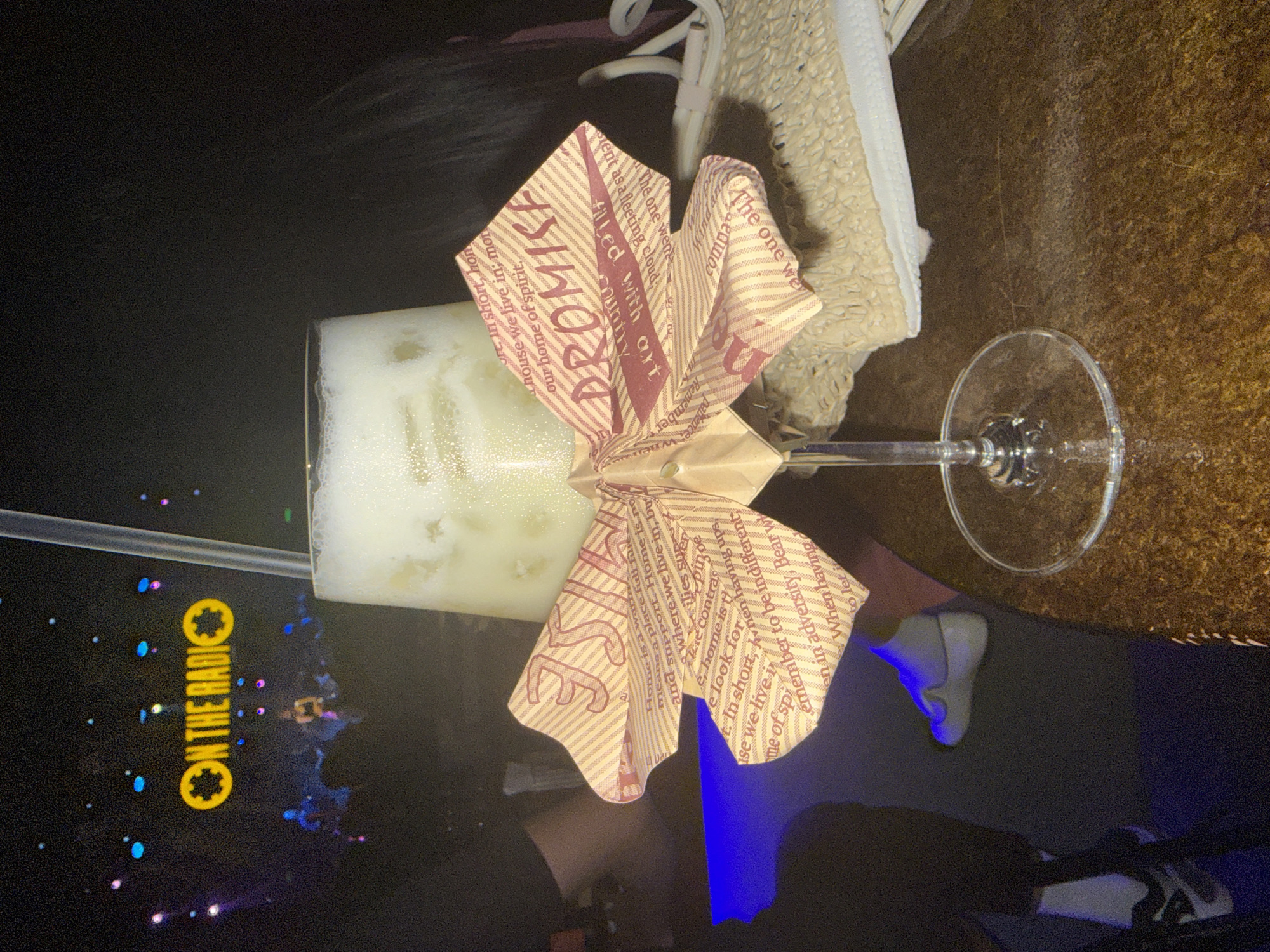 A rotated eye-level shot depicts a cocktail in a coupe glass adorned with a paper bow, a straw, and the corner of a sneaker. The cocktail is a light-colored, frothy concoction. The paper bow, predominantly red and tan, is imprinted with the word "Promise" and other text phrases. Adjacent to the glass is the corner of a beige and white sneaker with a textured surface and a white sole. A clear straw extends into the cocktail. The table surface is black, reflecting the bar's ambient lighting with the words "ON THE RADIO". There is a reflection of another person in the table. The table surface also shows a change to a dark brown wood surface to the top right of the frame.