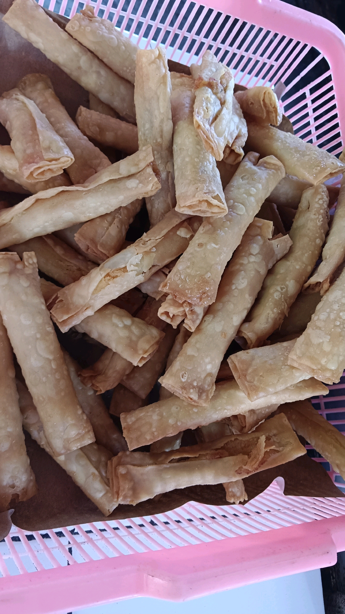A close-up, overhead shot captures a pile of golden-brown, fried spring rolls or similar snacks overflowing from a pink plastic basket. The snacks are cylindrical and appear to be made of thinly rolled dough, some with visible filling peeking through. The texture is crispy, with some pieces showing slight charring or browning. The background consists of the pink, slatted rim of the basket and dark, indistinct areas. The scene is well-lit, highlighting the texture and color of the food. The context of Jogorogo, Indonesia, suggests this could be a common street food or market scene.
