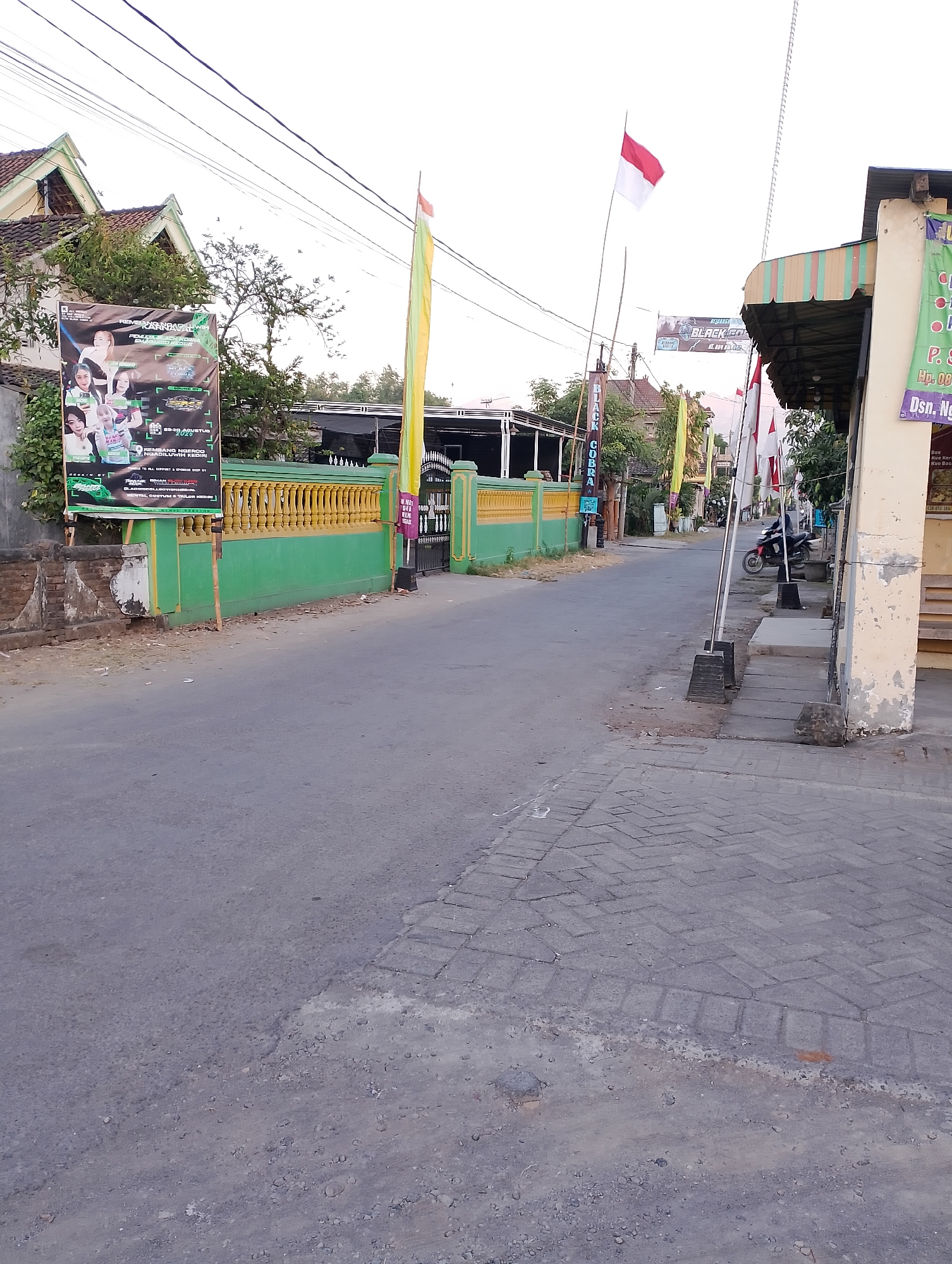 The image depicts a street scene in Rembang, Indonesia, likely in a residential or commercial area. In the foreground, a paved road transitions to a brick walkway. To the left, a bright green fence with yellow trim runs along the property line, bordered by a grey wall and a large banner advertising an event. A tall flagpole with an Indonesian flag stands near the banner. Further down the street, a covered structure with a striped awning is visible, with another Indonesian flag and more banners displayed. A red and black motorcycle is parked near the structure. The sky is overcast, and the overall lighting suggests it is either early morning or late afternoon. The buildings lining the street have traditional Indonesian architectural elements, with tiled roofs and simple facades. The street appears relatively quiet, with no people visible in the immediate vicinity.
