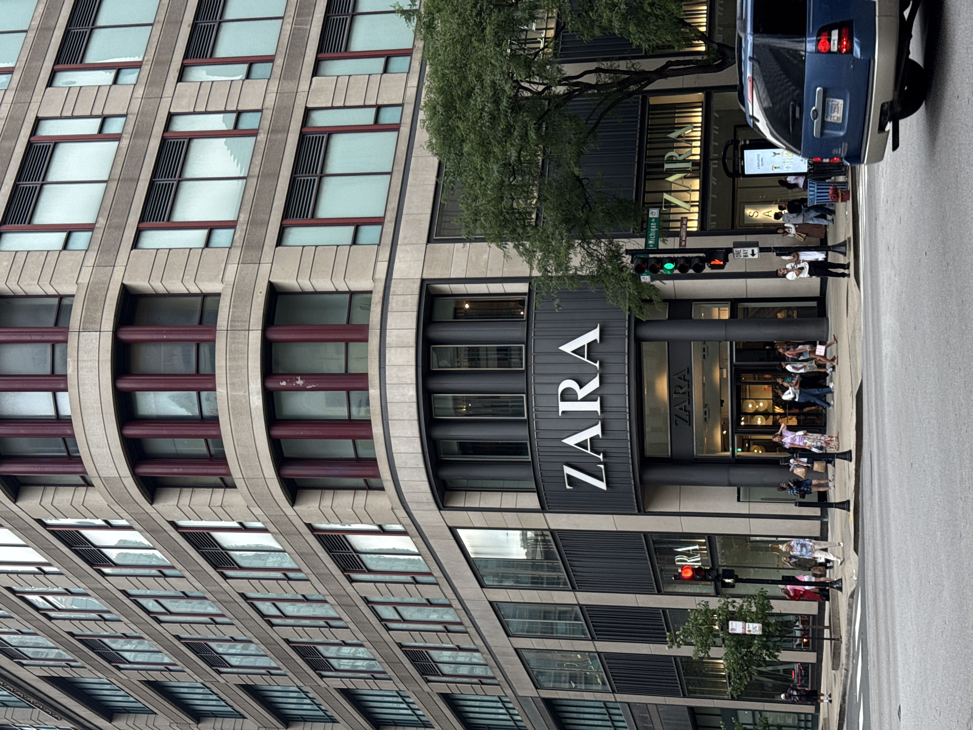 The image shows a Zara store located on Michigan Avenue in Chicago, Illinois, United States of America. The store is situated on the corner of a large, modern building with a beige stone facade and numerous windows. The Zara signage is prominently displayed in large white letters on a dark gray background above the store entrance.
Several people are seen on the sidewalk in front of the store, some are walking, while others appear to be browsing or waiting. A blue SUV is parked in the foreground on the street, obscuring part of the view of the store's lower level. Traffic lights and street signs are visible, indicating an urban street intersection. Green trees are also present along the sidewalk, providing some greenery amidst the urban architecture. The overall scene depicts a bustling commercial area in a city.