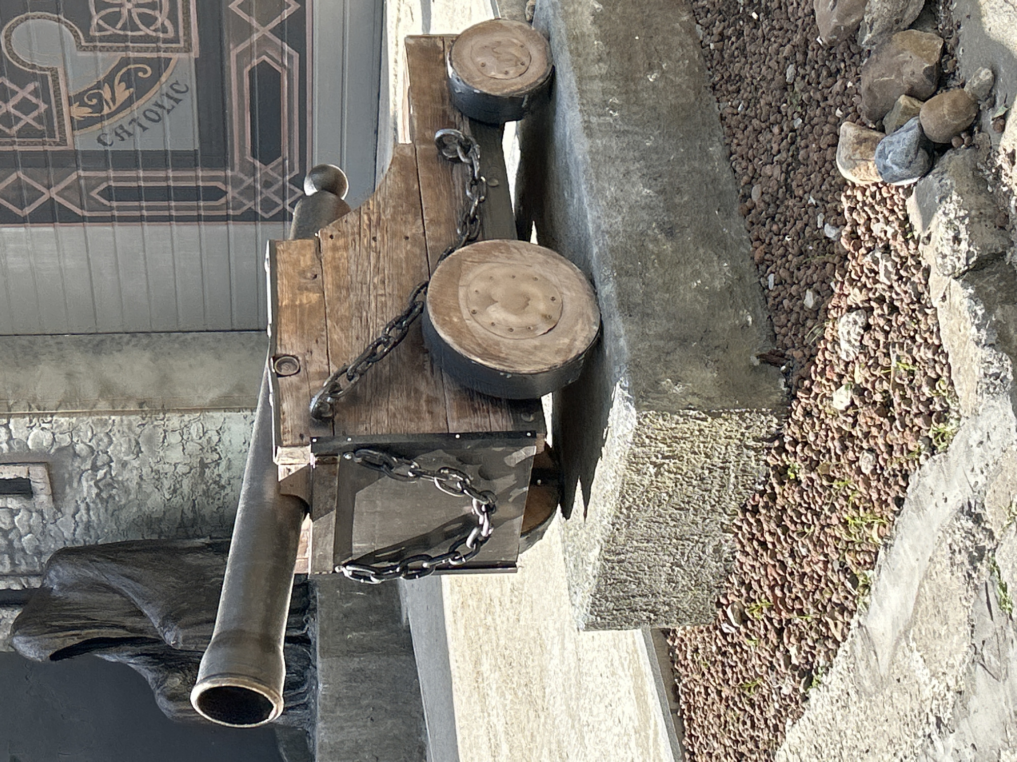 The image shows a close-up, eye-level view of a weathered wooden cannon replica placed on a stone pedestal. The cannon's barrel is pointed to the left, and it is supported by a wooden carriage with two large wooden wheels on each side, one at the front and one at the rear. Heavy chains are attached to the carriage. The cannon is situated outdoors, with a textured gray stone wall and part of a large metal door featuring ornate designs and the word "CATHOLIC" visible in the background. To the left of the cannon, a dark, hooded statue, possibly representing a cloaked figure, is partially visible. The ground around the pedestal is covered with small, reddish-brown pebbles. The scene suggests an architectural or artistic display. There is no indication that this image was taken in the city of Perm, Russia.