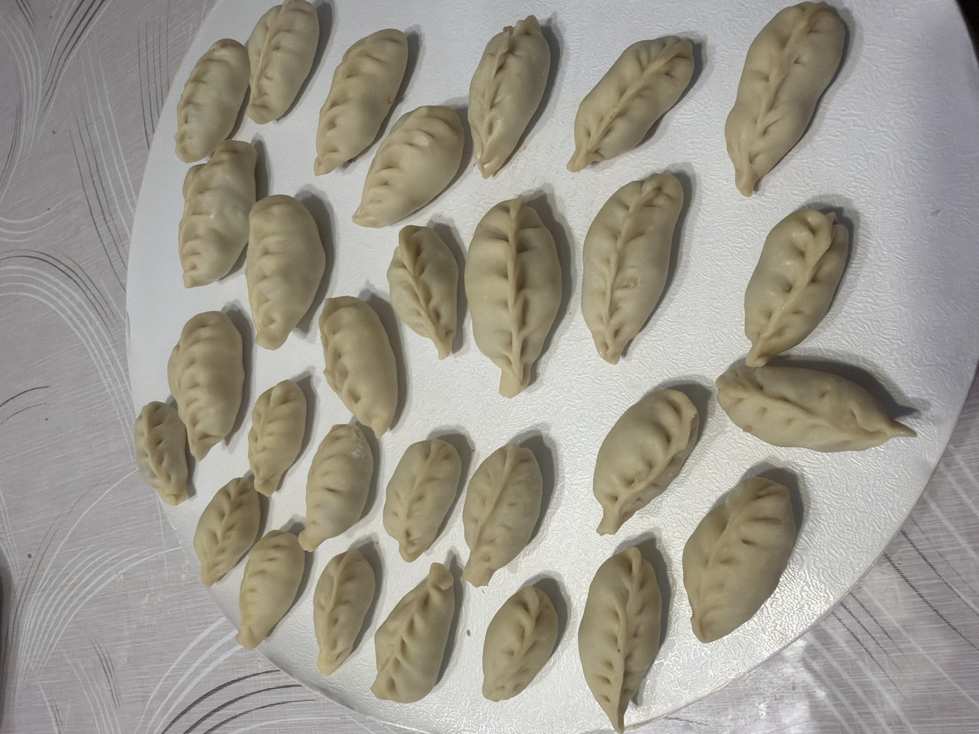 The image displays a white, textured circular surface, possibly a platter or tray, covered with numerous uncooked dumplings. The dumplings are arranged in rows and scattered across the surface. Each dumpling is pale yellow, doughy, and has a folded, pleat-like pattern along one side, resembling a crescent or a plump seed. Some dumplings are positioned closer to the viewer, while others recede into the background. The surface they rest on is situated on a table with a light-colored, patterned tablecloth underneath, featuring thin, wavy grey lines. The lighting suggests an indoor setting, likely a kitchen or dining area. The image appears to be a photograph taken from a slightly elevated angle, focusing on the dumplings. There are no people, animals, or any other objects visible in the frame, making the dumplings the sole subject of the photograph. The location is identified as Vernon Hills, United States of America, though no specific landmarks or contextual clues within the image confirm this.