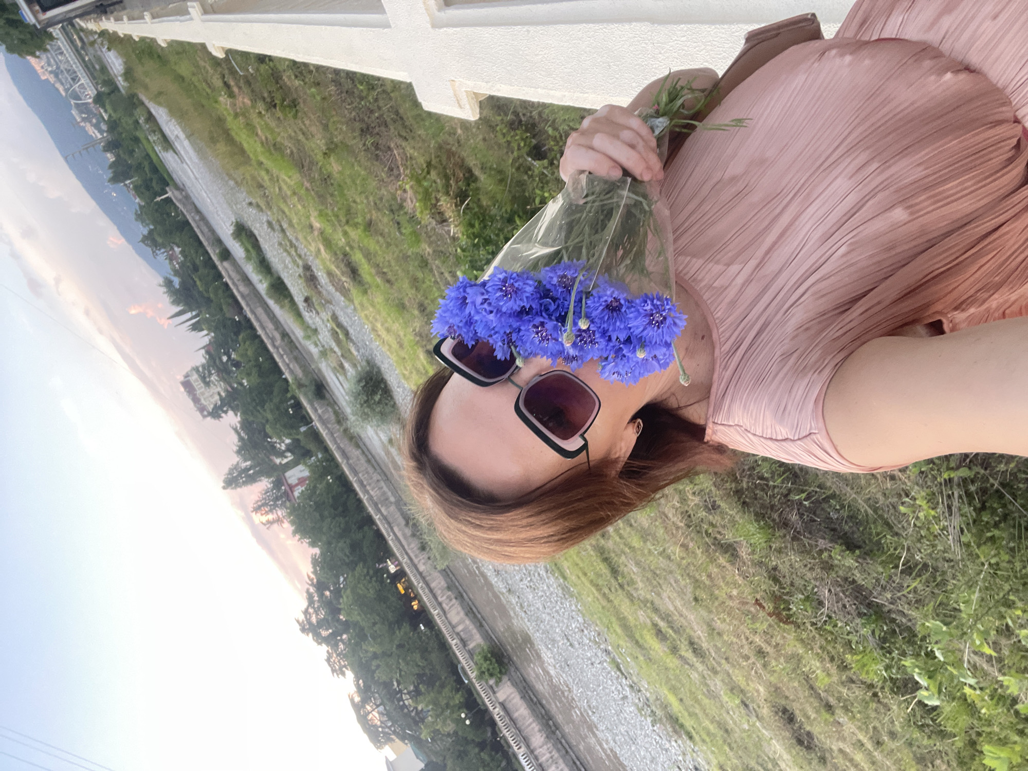 A woman with brown hair, wearing a pink dress and sunglasses, holds a bouquet of blue flowers in front of her face, obscuring her mouth and nose. She is standing on a paved area next to a white railing. In the background, a rocky riverbed is visible, with a river flowing through it. Trees and buildings line the far bank of the river, with mountains in the distance. The sky is overcast with some pink hues suggesting it might be near sunset. The scene is likely in Sochi, Russia, given the presence of a river, mountains, and lush greenery typical of the region.