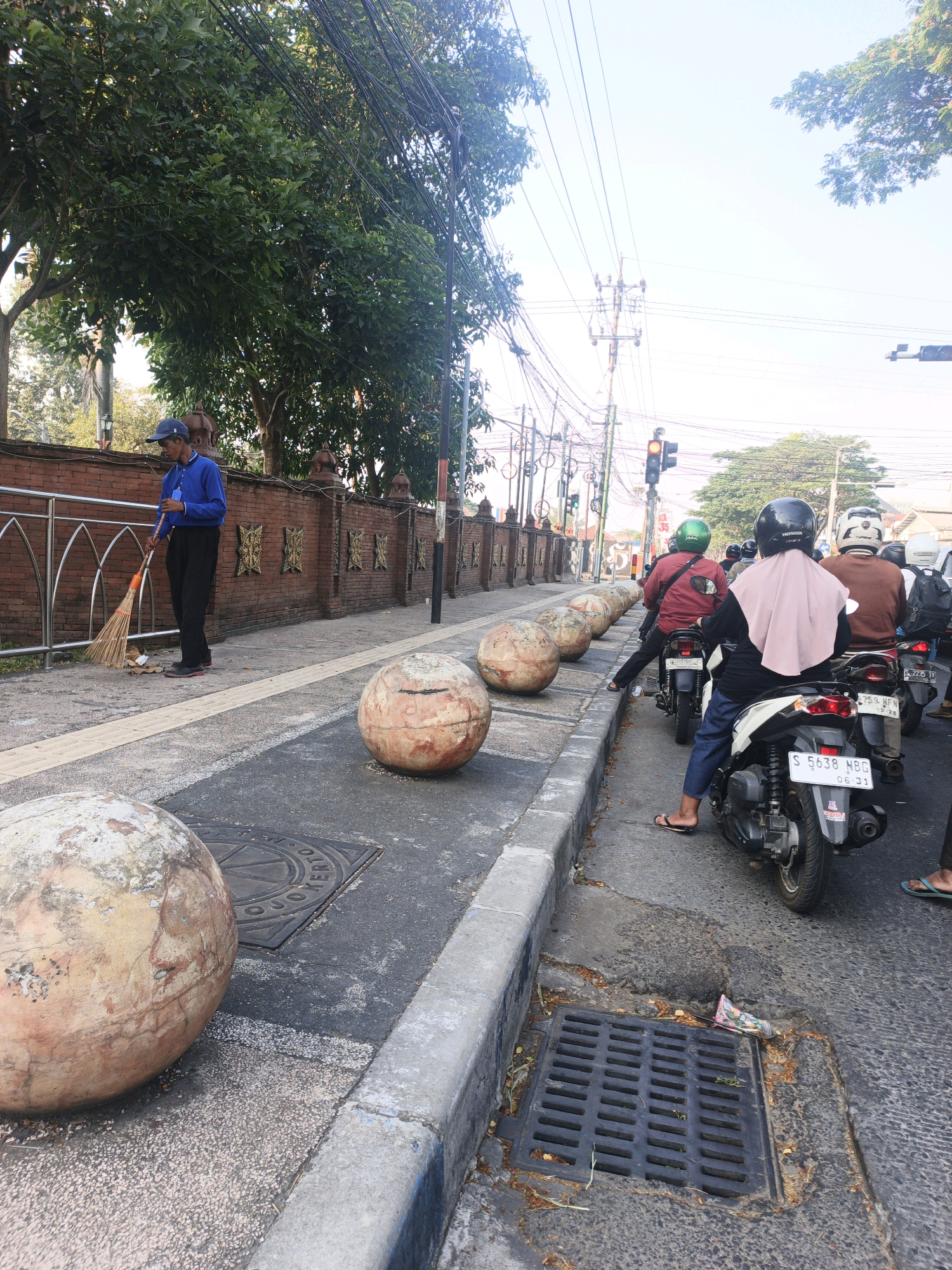 The image depicts a street scene in Sooko, Indonesia. A sanitation worker wearing a blue shirt and a blue cap is sweeping the sidewalk with a broom. The sidewalk is bordered by a brick wall with decorative patterns and a metal fence. Large, spherical concrete bollards are placed along the sidewalk. In the background, several motorcycles are stopped at a traffic light, with riders wearing helmets. The street is lined with trees and overhead utility wires are visible. The scene suggests an urban environment with daily activities taking place.