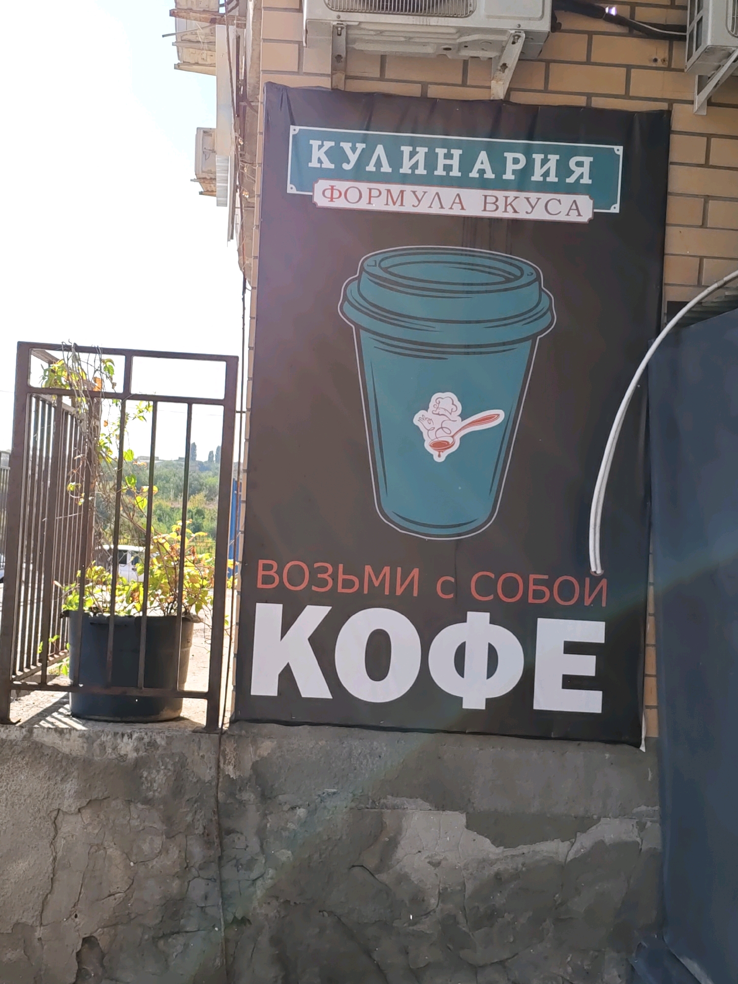 The image shows a large banner with Cyrillic text and a graphic of a coffee cup. The banner is mounted on a brick wall and appears to be advertising a cafe or coffee shop. The text on the banner reads "КУЛИНАРИЯ" (Culinary) and "ФОРМУЛА ВКУСА" (Formula of Taste) at the top, with a large graphic of a teal coffee cup below. At the bottom, the text reads "ВОЗЬМИ С СОБОЙ КОФЕ" (Take Coffee With You). The banner is partially obscured by a metal fence and a potted plant. The environment suggests an outdoor setting, possibly on a street or walkway, with a glimpse of greenery and a cloudy sky in the background. This scene is consistent with a commercial establishment in Rostov-on-Don, Russia.