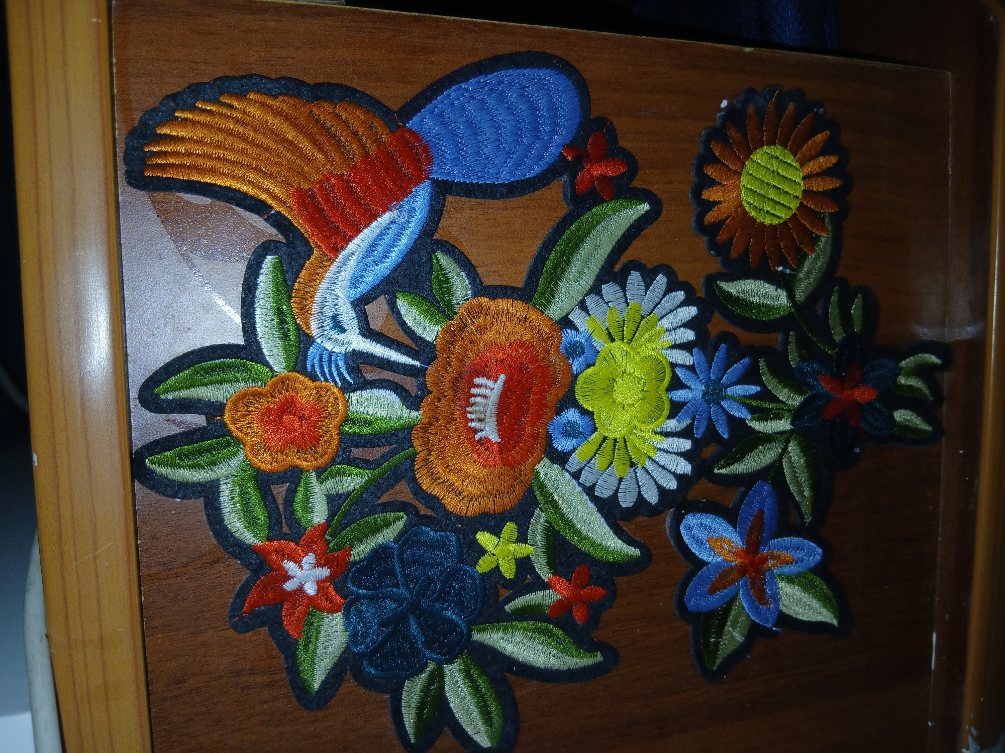 This is a close-up, eye-level photograph of an embroidered appliqué featuring a bird and flowers, affixed to a wooden surface. The appliqué is primarily composed of vibrant thread work. A bird with an orange, red, and blue body and wing is positioned at the top right, partially behind a cluster of stylized flowers. The flowers vary in color, including red, orange, blue, yellow, and white, and are rendered with intricate petal details. Green leaves with black outlines surround the floral elements. The wooden surface appears to be part of a piece of furniture, possibly a cabinet or dresser, with a visible wood grain and a slightly glossy finish. The lighting is moderate, casting subtle shadows. The photograph does not contain any elements that would place it in the city of Тайгараев, Kyrgyzstan.