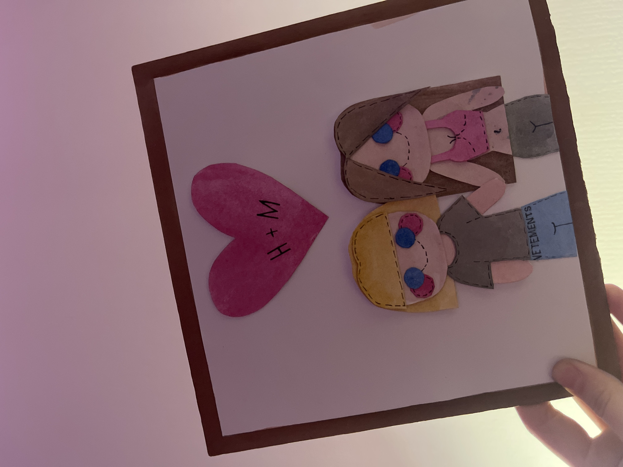 The image shows a framed piece of artwork held in someone's hand. The artwork is made from felt or similar material and depicts two figures and a heart. One figure has brown hair and wears a pink top and gray shorts. The other figure has blonde hair, a gray shirt with the word "VETEMENTS" printed on it, and blue shorts. A pink heart shape with the letters "H+M" written on it is also present on the white background. The frame is brown. It is not possible to determine the location within Saint Petersburg, Russia, from the provided image.
